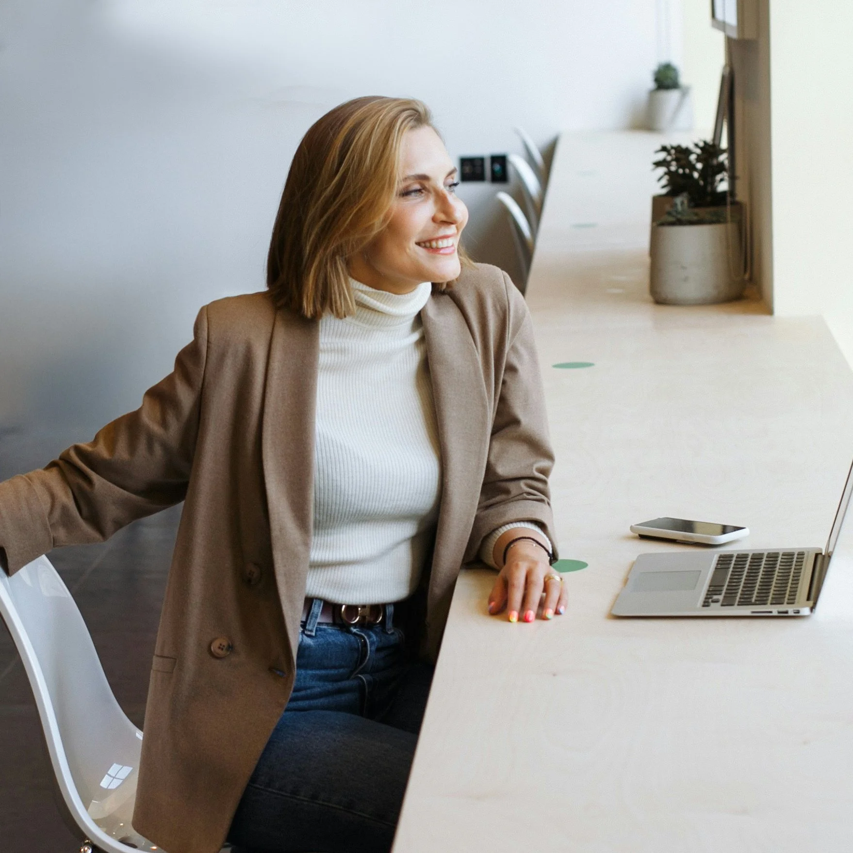 Woman with short blonde hair in a tan blazer and white turtleneck staring out a window and smiling