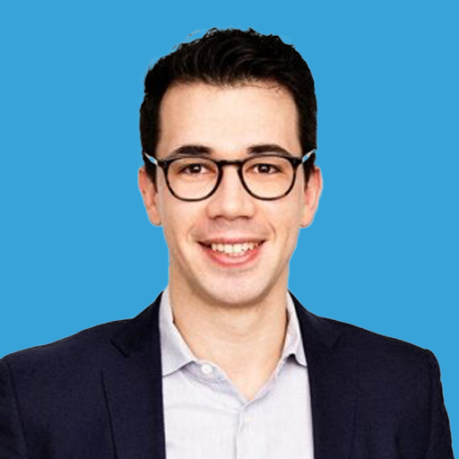 A smiling young man with black hair, glasses, and fair skin wearing a navy blazer and light shirt against a blue background.
