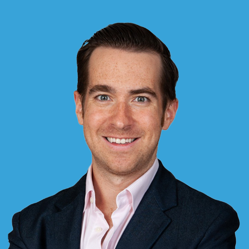 Portrait of a smiling man with dark hair, wearing a navy blazer and a white shirt, against a blue background.
