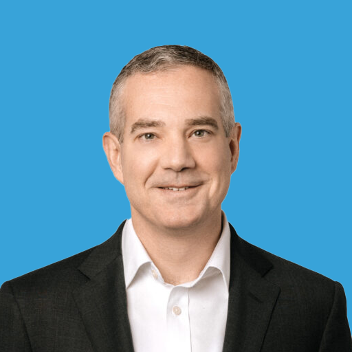 Headshot of a man with short gray hair, wearing a white shirt and black suit jacket, smiling against a blue background.