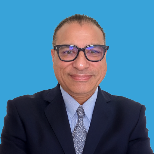 A man wearing glasses, a dark suit, light blue shirt, and patterned tie, smiling against a solid blue background.