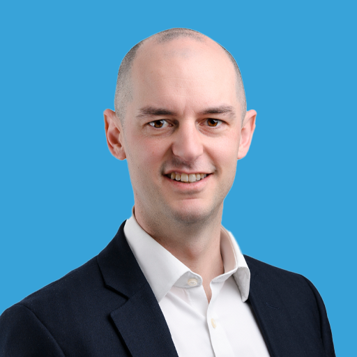 A professional portrait of a man with a shaved head wearing a white shirt and a dark blazer, smiling against a blue background.
