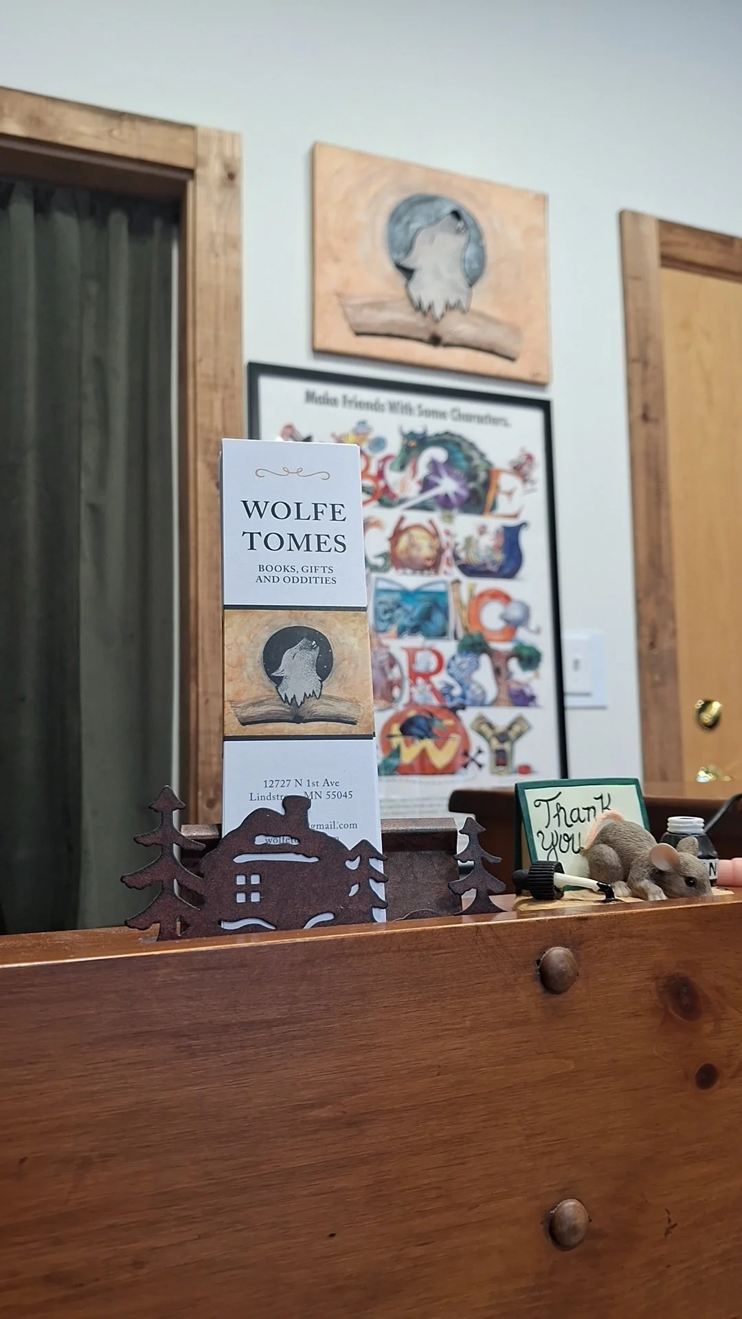 Bookmark for Wolfe Tomes with logo of a wolf's head, placed on a wooden surface along with a small mouse figurine and a 'Thank You' sign. Background features a framed cartoon artwork and a wall decoration with a wolf's head.