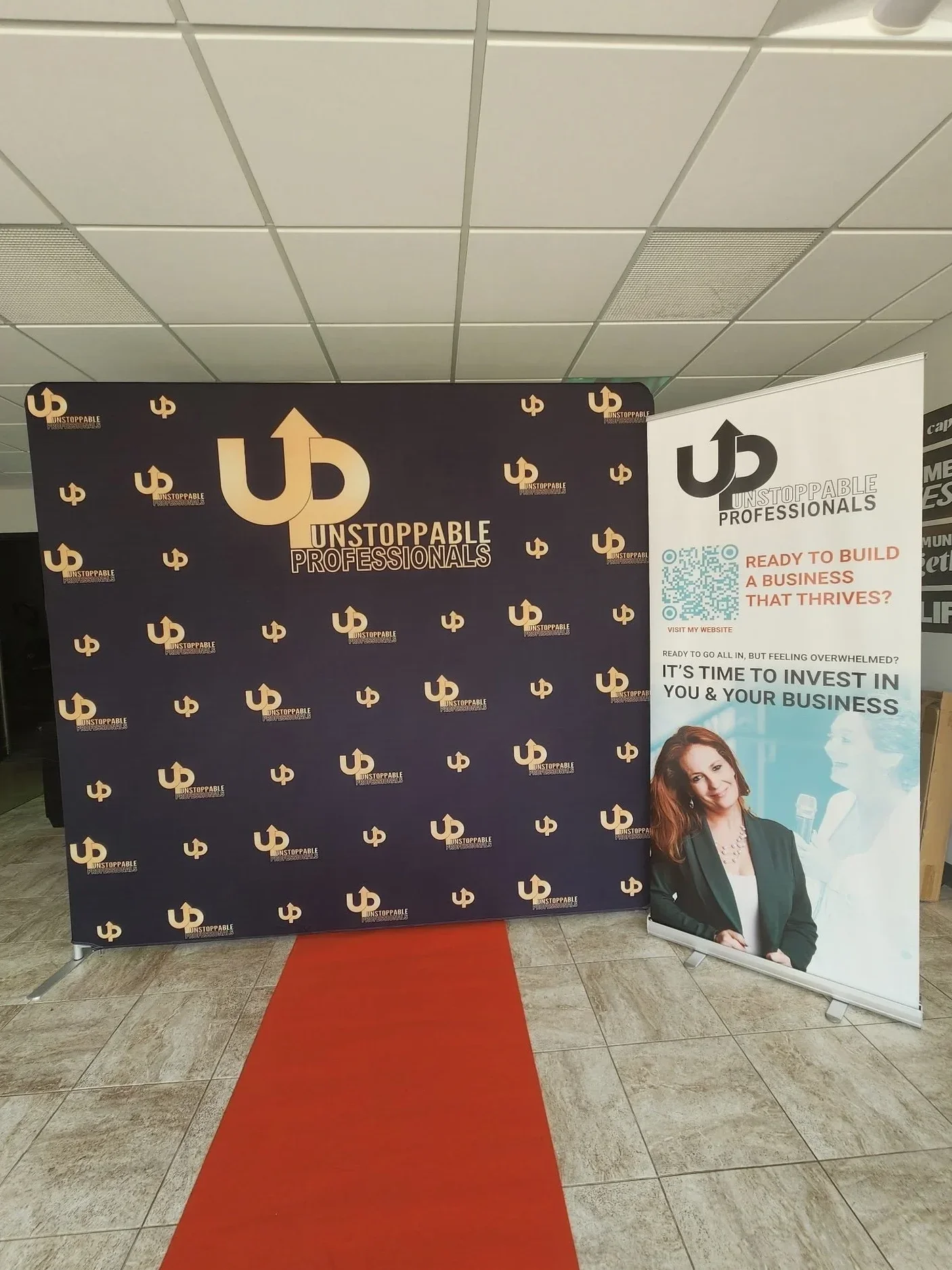 A black backdrop with multiple repeating logos and text that reads 'Unstoppable Professionals'. Beside the backdrop, there is a standing banner with the same logo and additional text about building a successful business. The banner shows a smiling woman with curly red hair, wearing a dark blazer, and there is a red carpet on the floor.