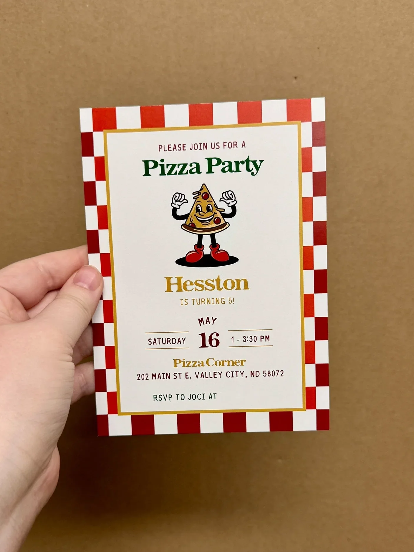 A person holding a pizza party invitation with a checkered red and white border, featuring a cartoon pizza slice with a smiling face and arms, and text detailing the event at Pizza Corner in Valley City, North Dakota.