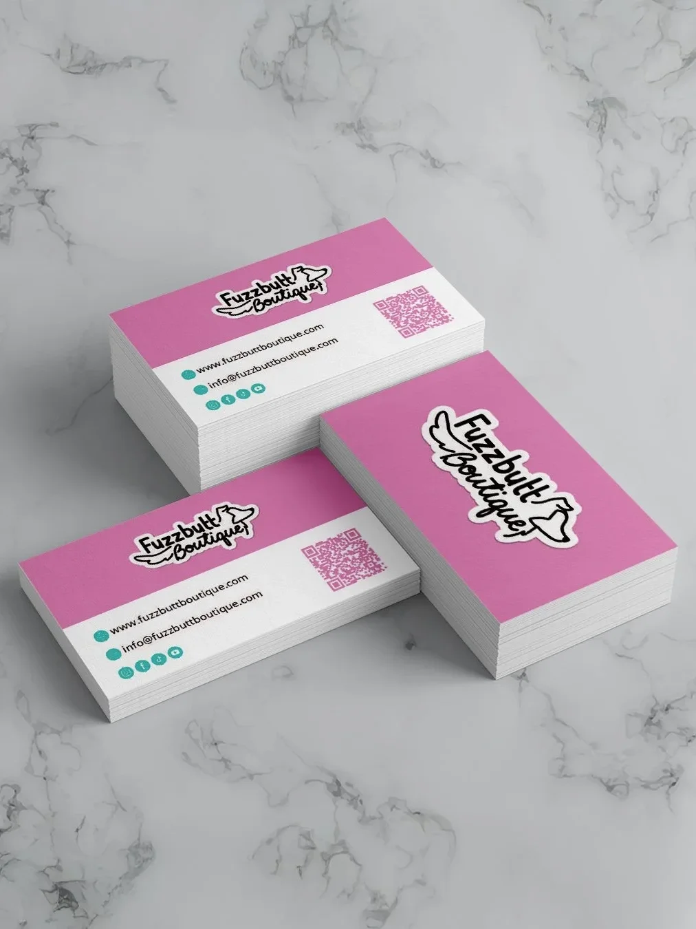 Stacks of business cards for FuzzButt Boutique with pink background, logo featuring a dog, contact information, and a QR code, on a marble surface.