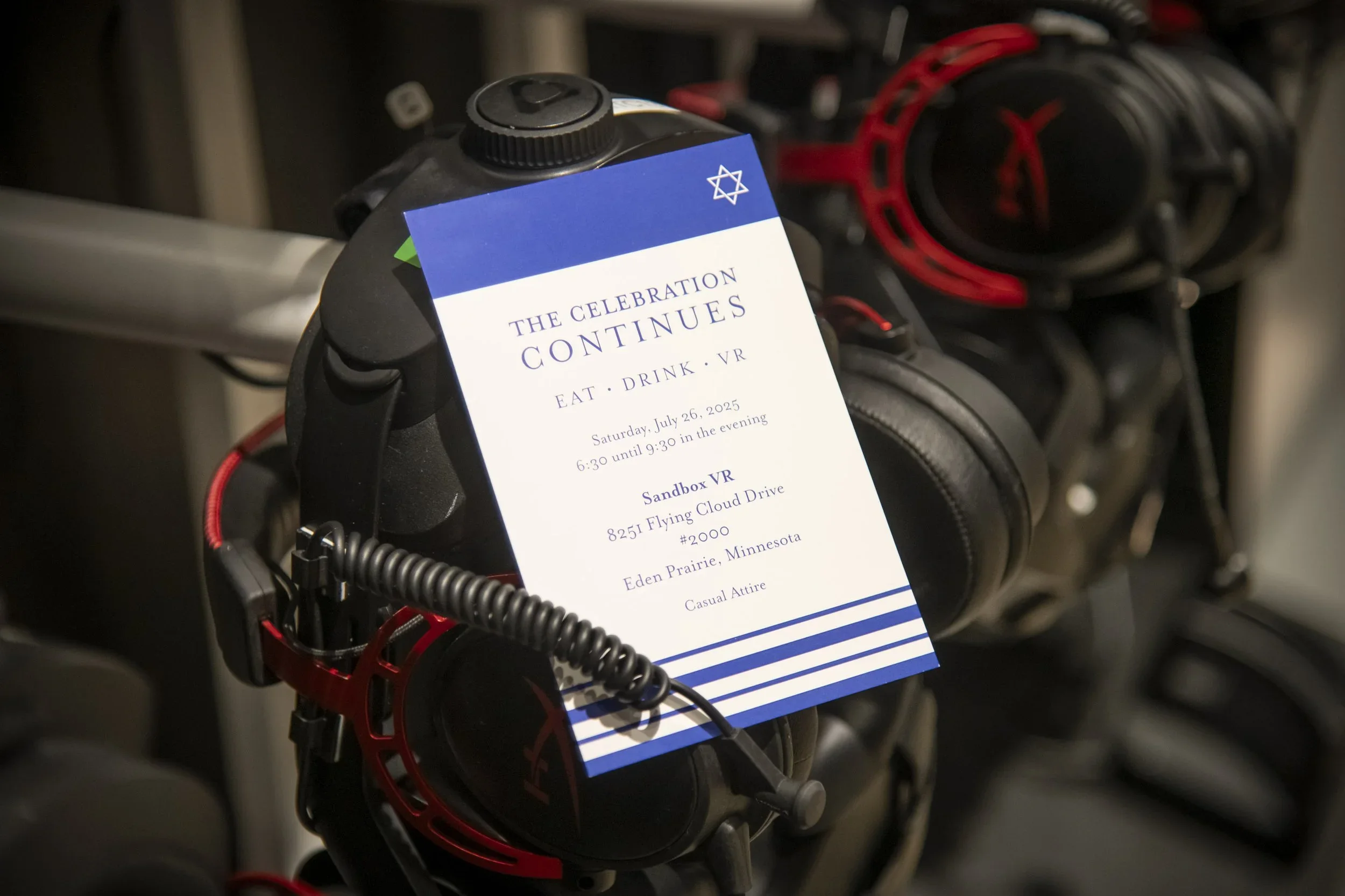 Close-up of a black device with red accents, with a blue and white event invitation attached that reads 'The Celebration Continues,' and includes details about a virtual event on July 26, 2025, in Eden Prairie, Minnesota.