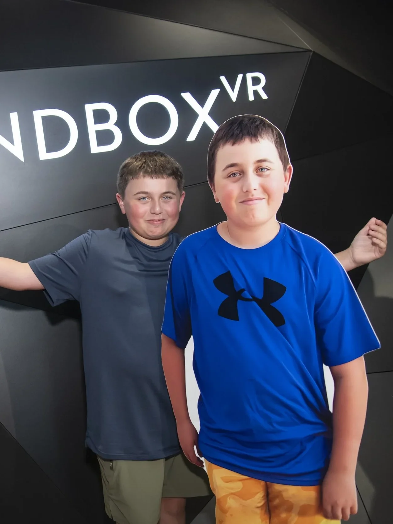 Two young boys standing in front of a black wall with the words 'NDBOX VR' illuminated. One boy wears a gray shirt and the other wears a blue shirt with a black Under Armour logo and orange shorts.