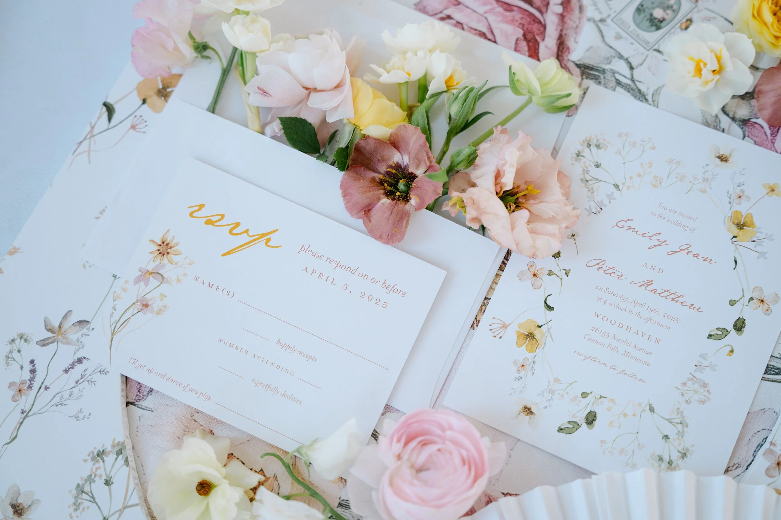 Wedding invitation cards with floral designs, surrounded by pink, white, and peach flowers on a table.