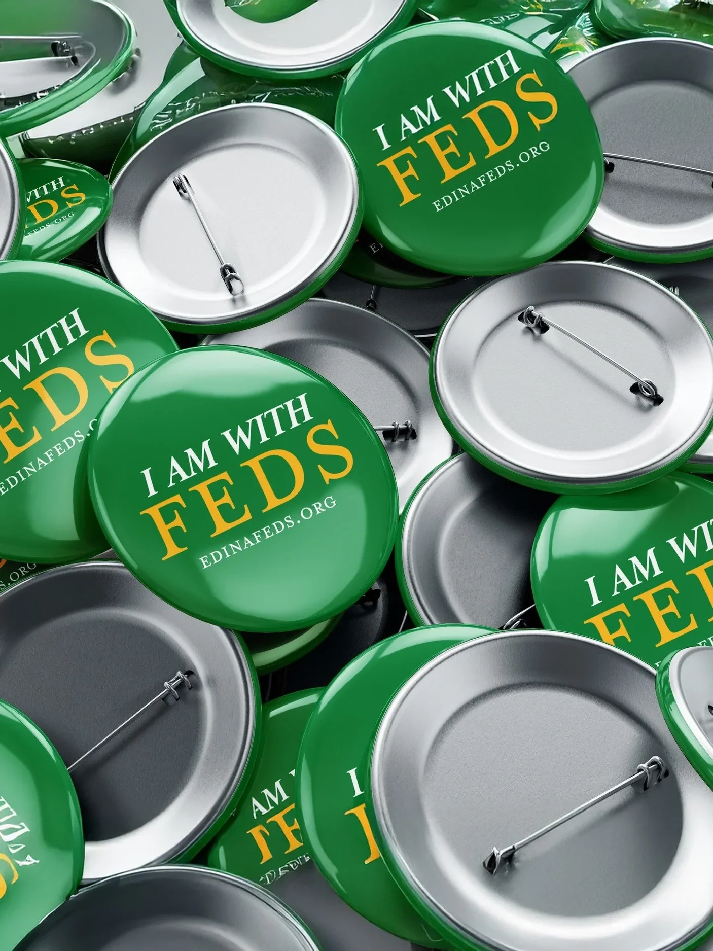 A pile of metal badges with green backs and safety pins, displaying the message "I AM WITH FEDS" and the website edinafeds.org.