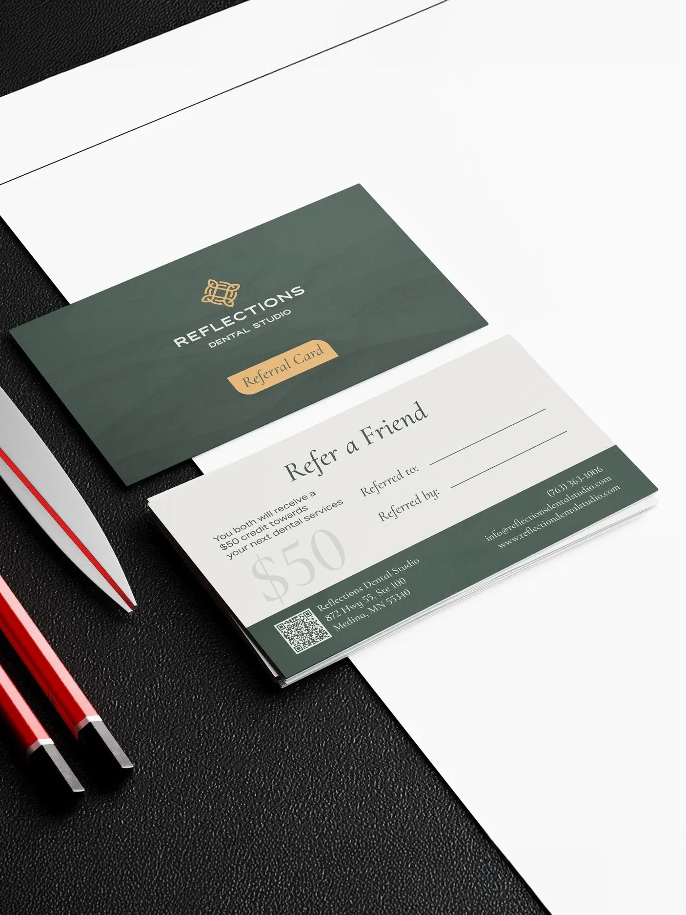 Referral card for Reflection Dental Studio, offering a $50 credit towards dental services, enclosed in a white envelope, on a black textured surface with two red and black pens nearby.