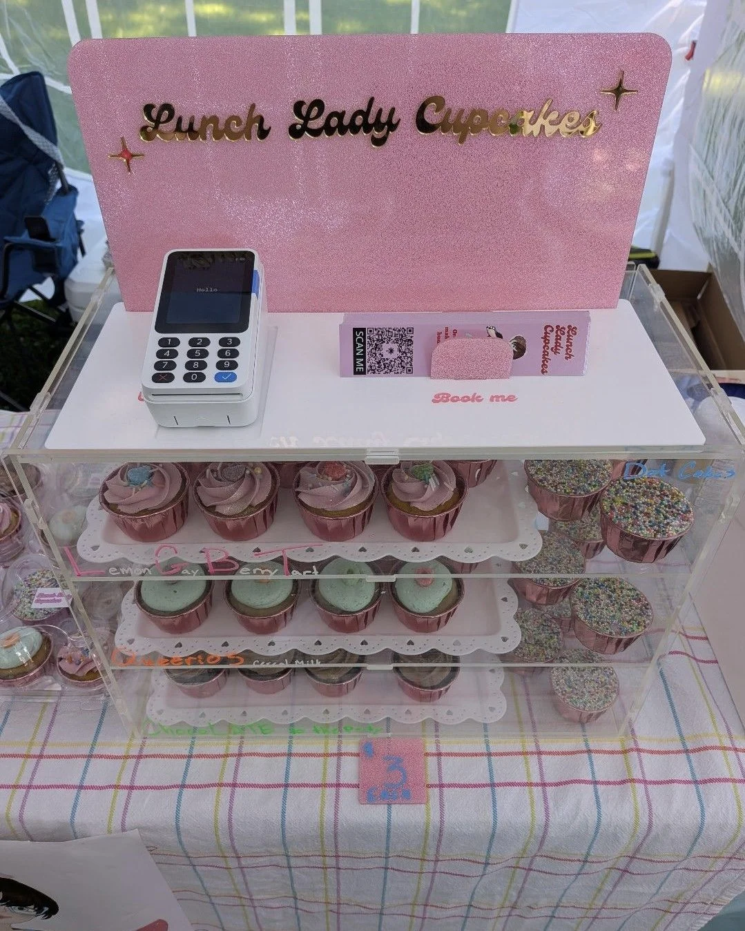 Display case with pink cupcakes topped with pastel frosting and colorful sprinkles, labeled 'Lemon', 'B', and others. Pink sign reading 'Lunch Lady Cupcakes,' with a small card and a point-of-sale device on a checkered tablecloth.