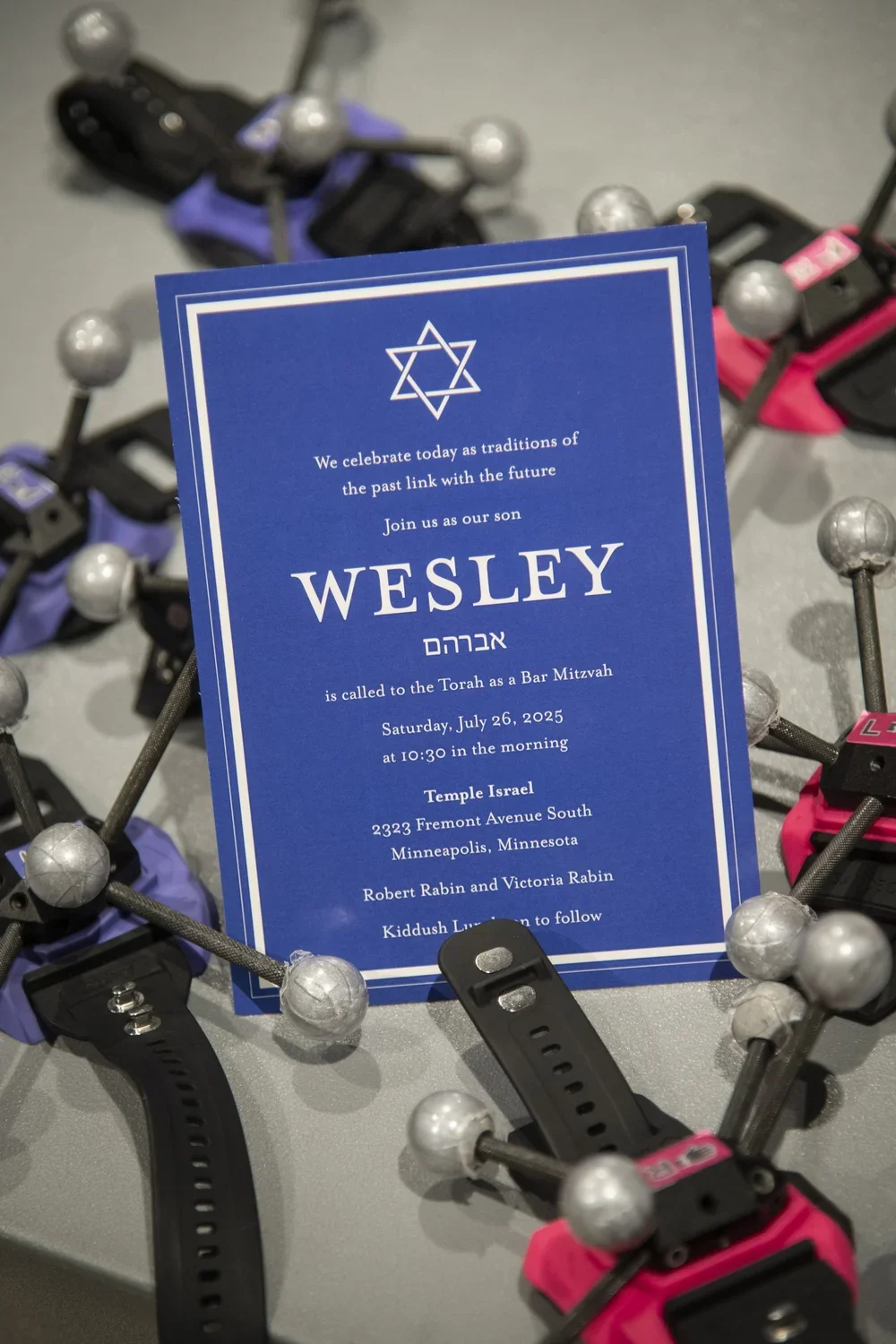 A blue Bar Mitzvah invitation card with a Star of David at the top, surrounded by foosball tables with pink and purple handles in the background.