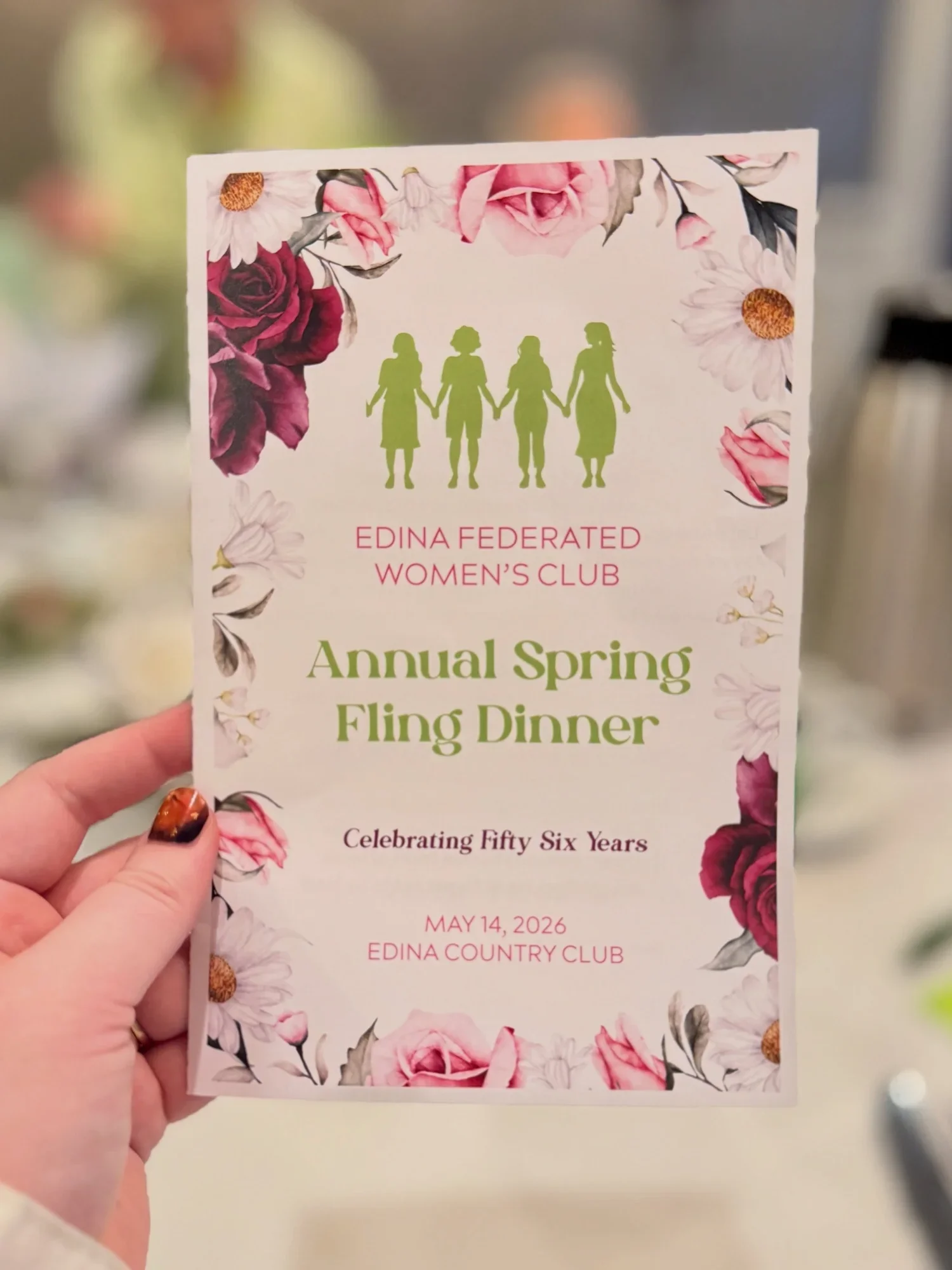 A woman holding a floral invitation card for the Edina Federated Women's Club Annual Spring Fling Dinner. The card has pink, white, and red roses and daisies with green leaves around the border. The event is scheduled for May 14, 2026, at the Edina Country Club.