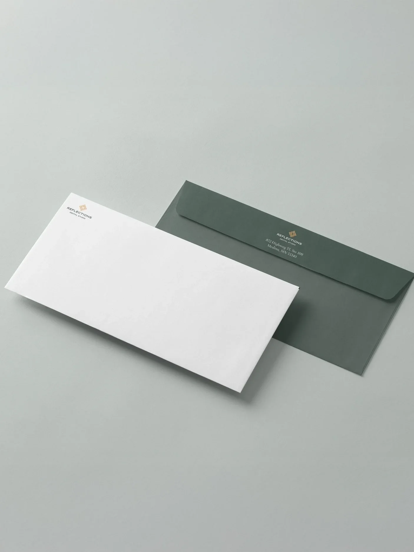 Two envelopes, one white and one dark green, on a light gray surface. The envelopes have the logo and address of a company named 'Reflections Dental Studio' printed on them.