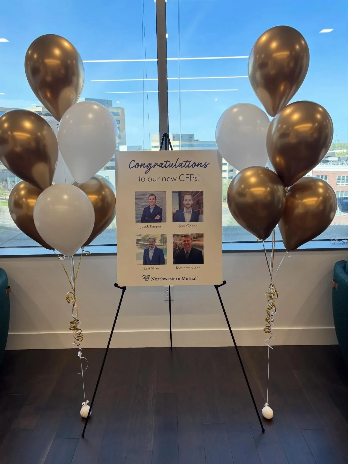 Balloon display with four black and three white latex balloons, all with curled ribbons, arranged around a sign on a stand. The sign congratulates new CFPs and features photos of four individuals with their names and the Northwestern Mutual logo. The background shows a large window with a cityscape and buildings outside.