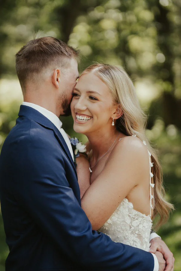 A newlywed couple sharing a joyful moment outdoors, with the bride smiling and wearing pearl earrings and a lace wedding dress, and the groom in a navy blue suit, in a lush green park setting.