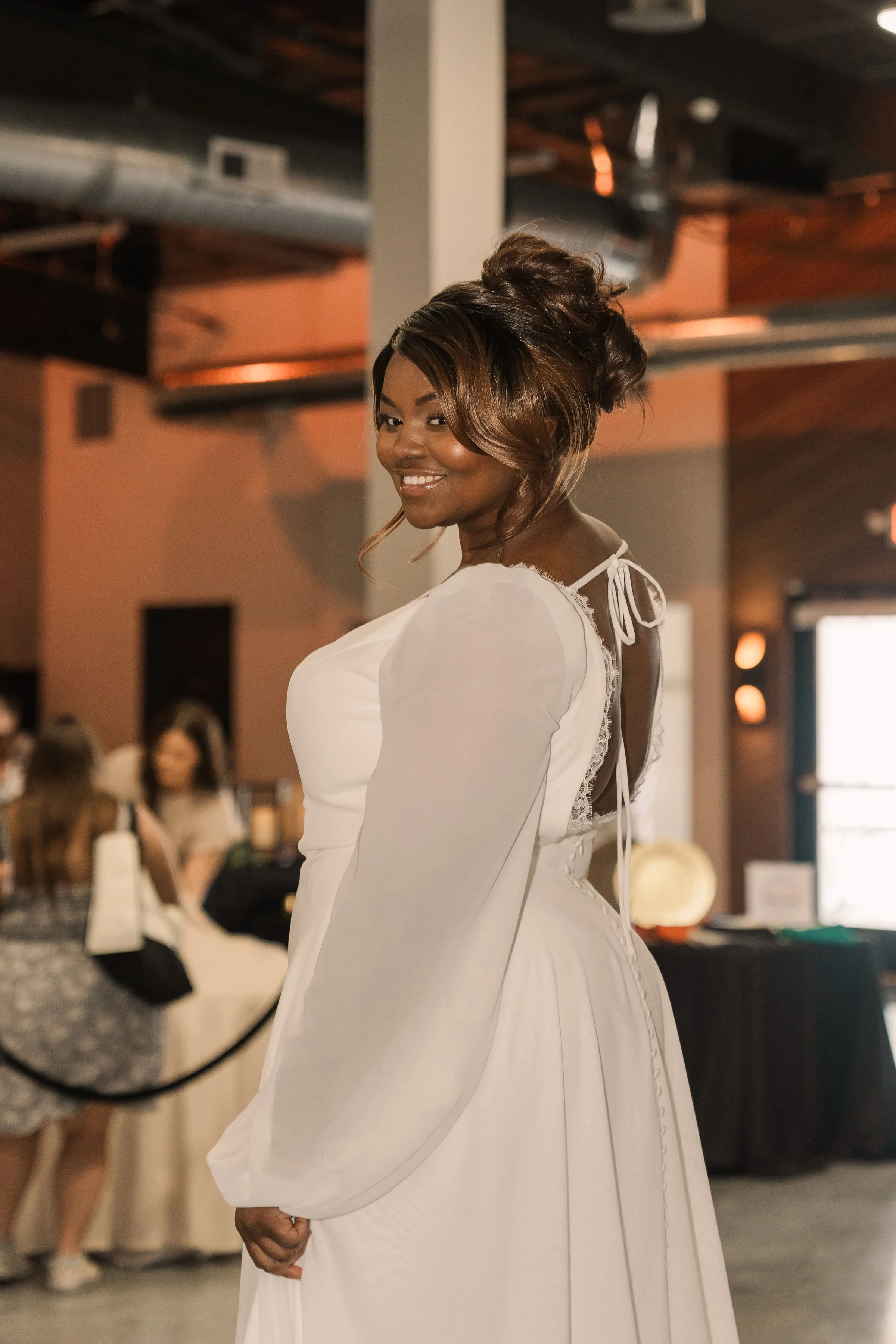 A woman in a white dress smiling at the camera in an indoor setting with people seated at tables in the background.