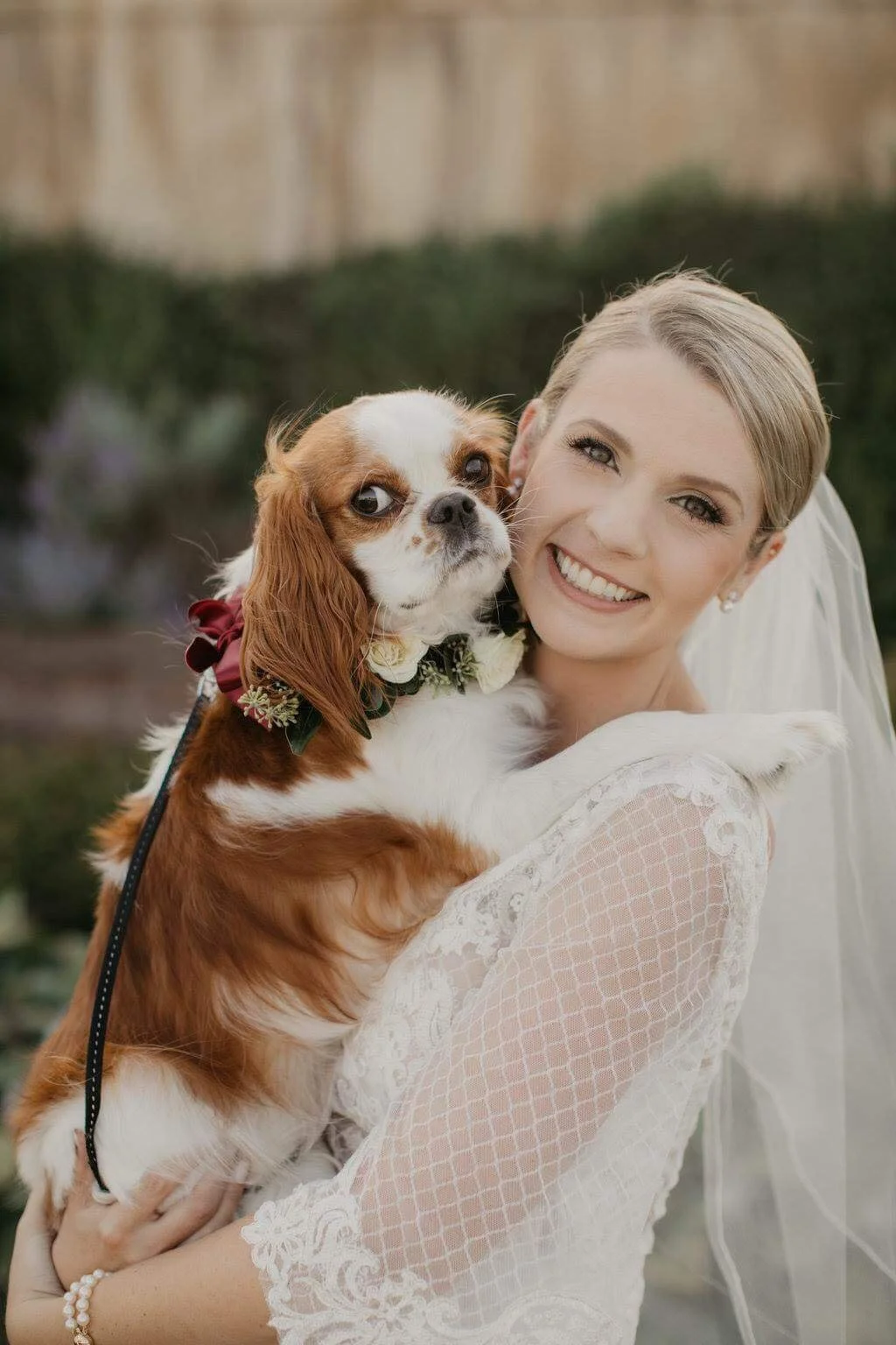 A bride smiling and holding a Cavalier King Charles Spaniel dog close to her face.