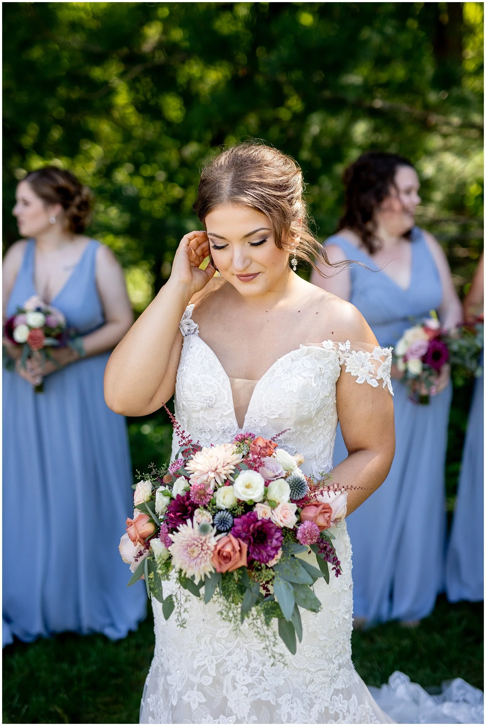 A bride in a lace wedding gown holding a colorful bouquet of flowers, with her eyes closed and hand touching her hair, surrounded by bridesmaids in blue dresses holding bouquets in an outdoor setting.