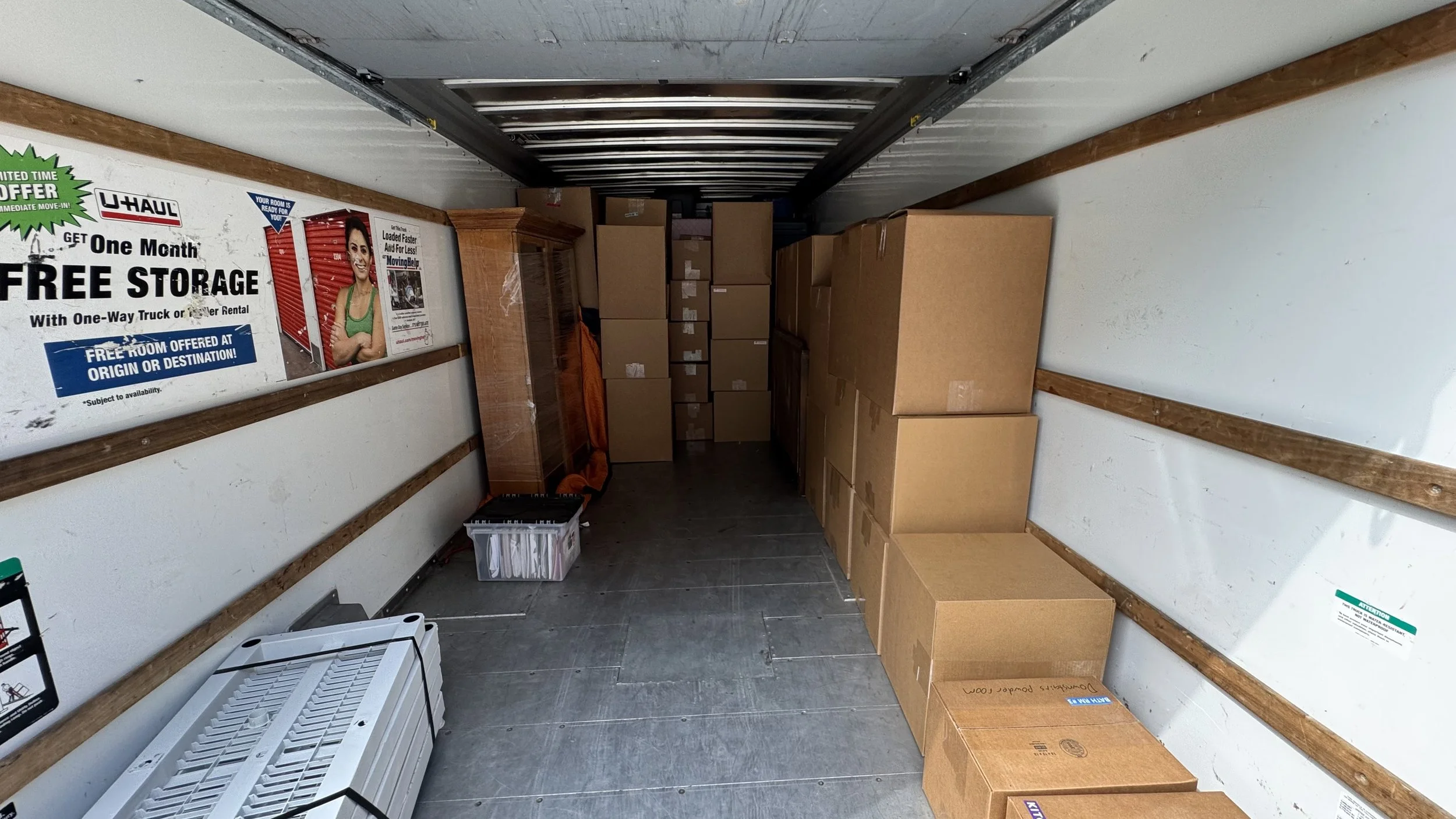 The interior of a moving truck filled with stacked cardboard boxes, a plastic storage bin, a small wooden cabinet, and a white appliance, with a U-Haul advertisement poster on the left wall.