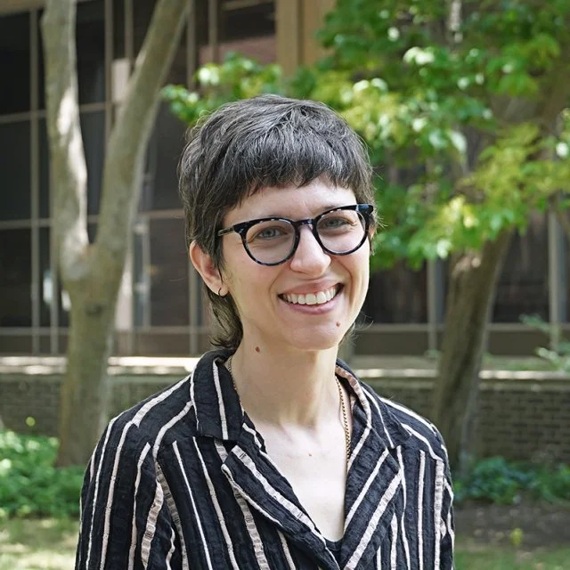 A woman with short black hair smiling outdoors, wearing a blue top and layered necklaces, with a background of green foliage and purple flowers.