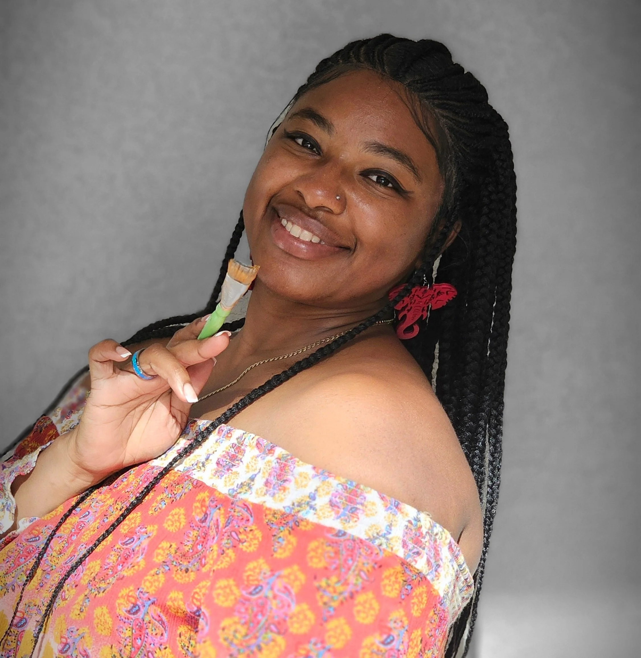 Latch and Paint founder and creator wearing braids, wearing colorful off-shoulder clothing and red earrings, smiling and holding a paintbrush against a gray background.
