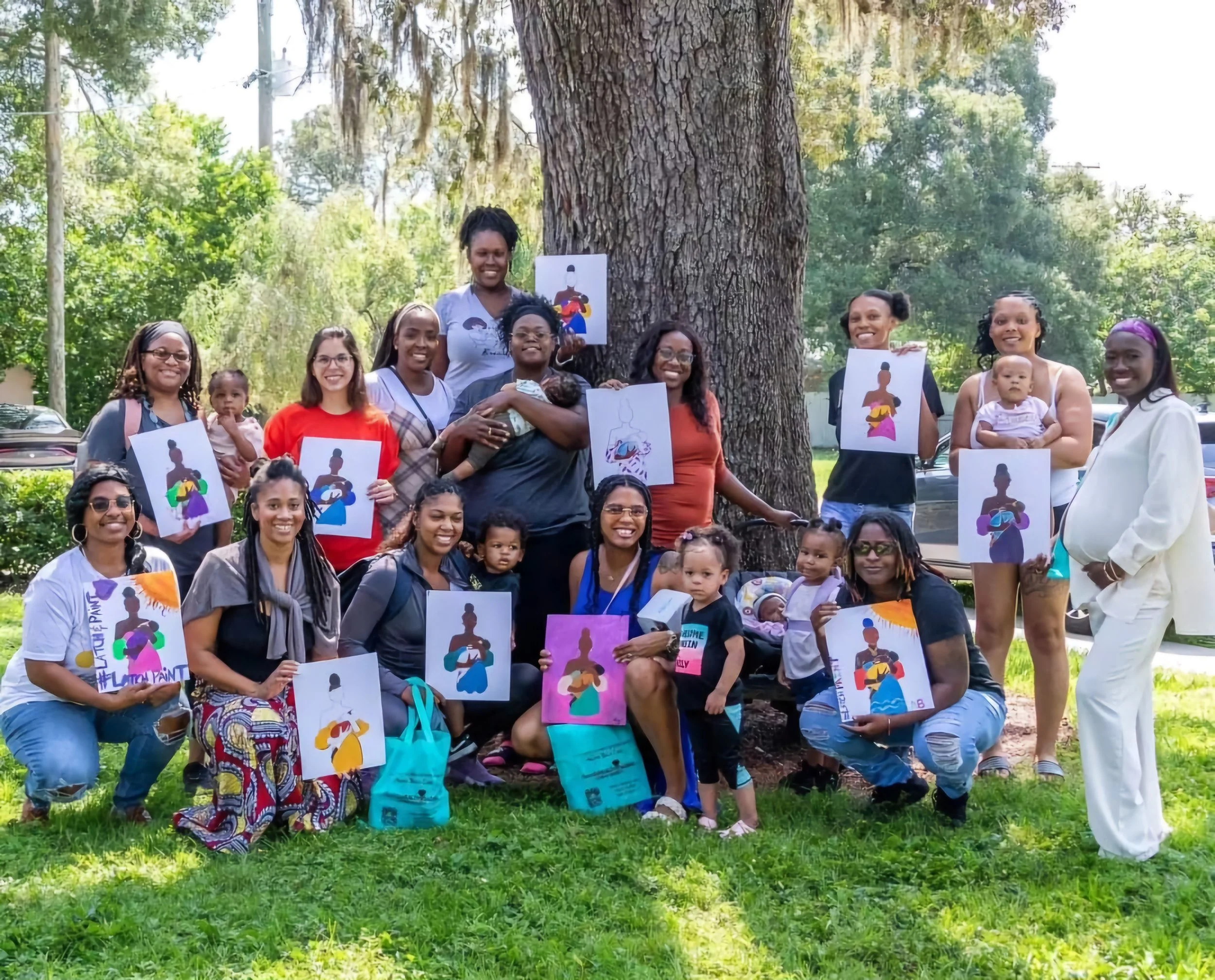 Group of women and children outdoors holding colorful art prints featuring a silhouette of a woman with a bun hairstyle, next to a large tree on a sunny day.