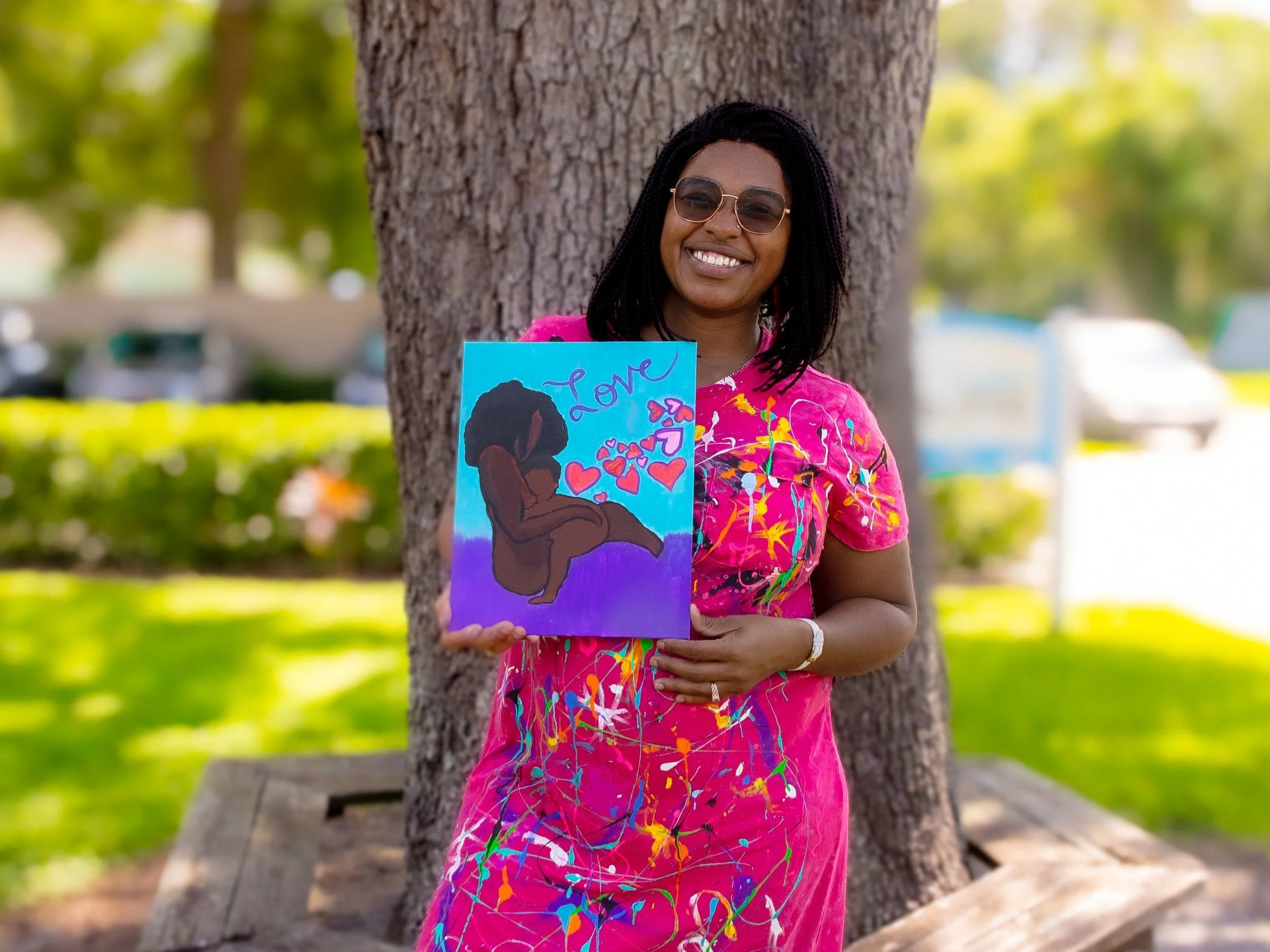 standing outdoors in front of a large tree, holding a colorful painting of a silhouette of a girl with the word 'Love' and hearts. She is wearing sunglasses and a vibrant pink dress with colorful splashes. A decorated sign with the message Hello The background features greenery and parked cars.