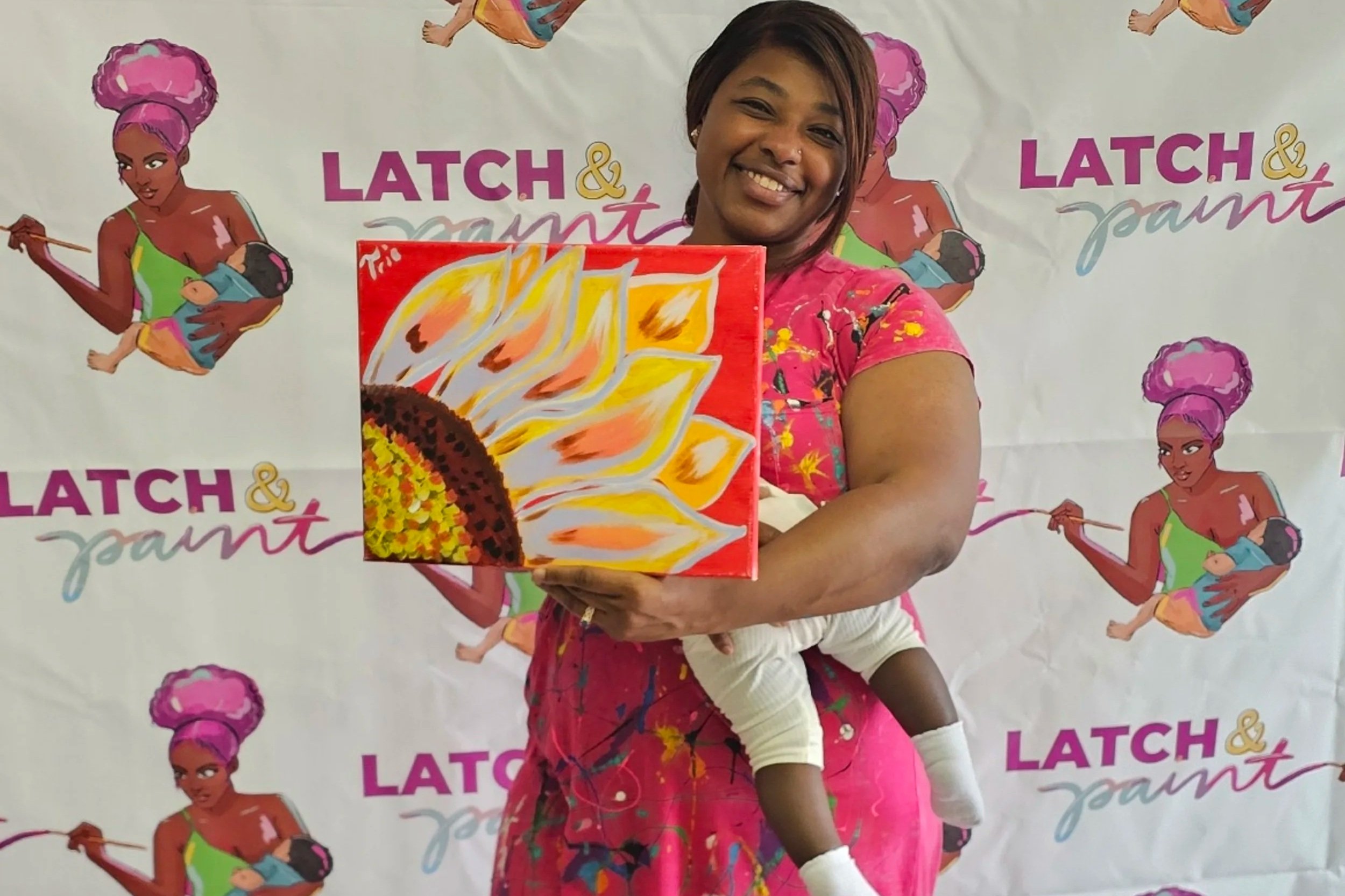 Founder and owne in a pink dress holds a painting featuring bright, abstract flames and a donut with sprinkles. She stands in front of a backdrop that reads 'LATCH & paint' and shows a logo of a woman with purple hair holding a paintbrush and a baby.