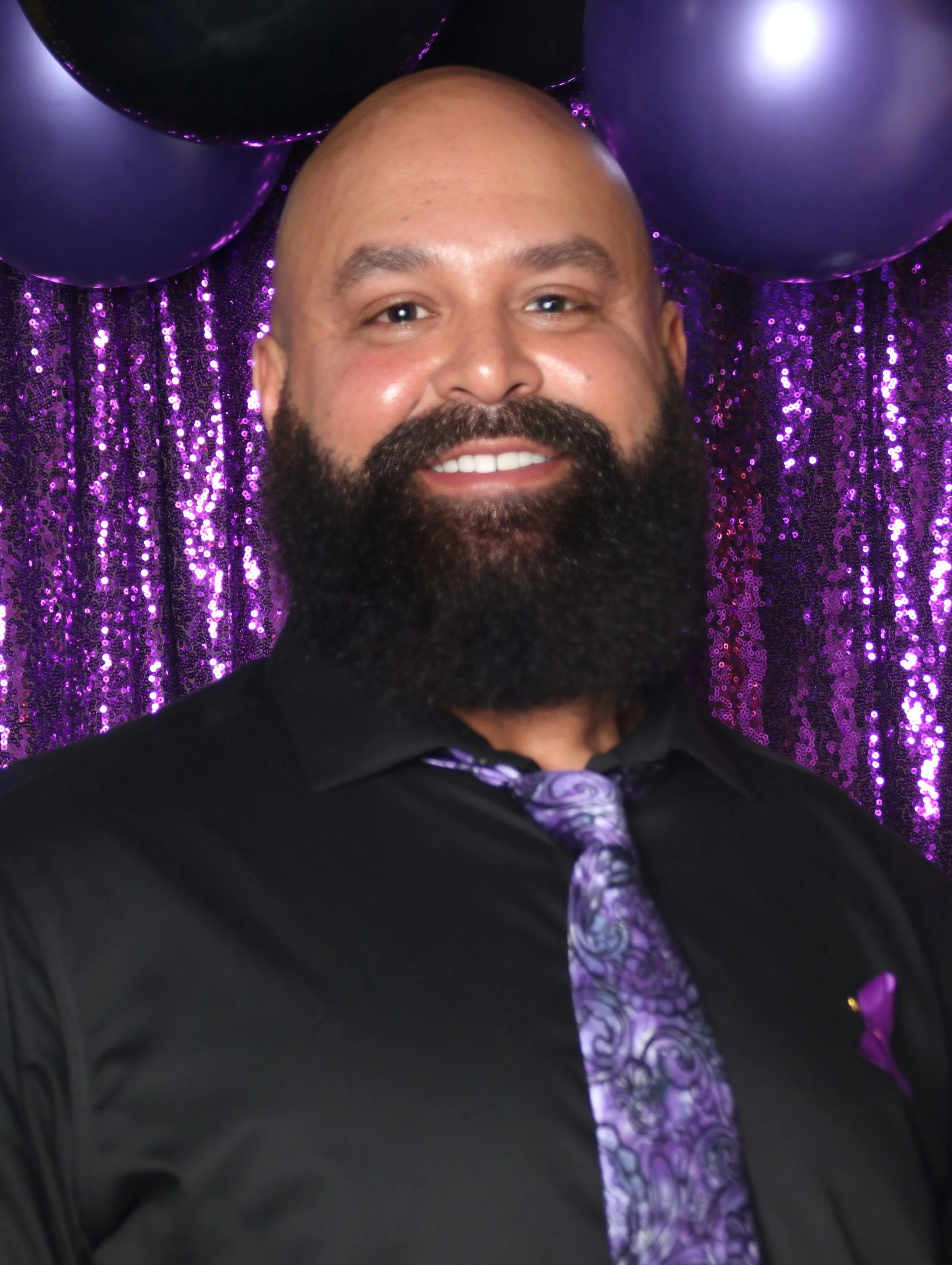 A man with a beard wearing a black shirt and patterned purple tie stands in front of a purple sequin backdrop with purple balloons.