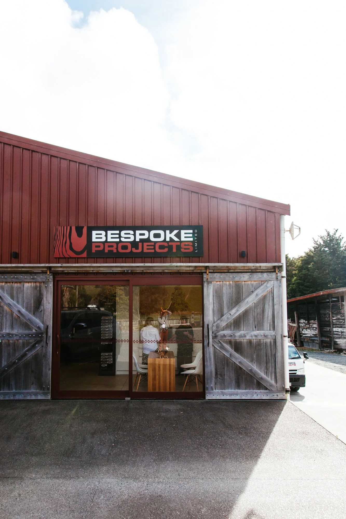 Front view of a red barn-style building with a sign that reads 'Bespoke Projects Limited'. The building has wooden sliding barn doors, with a glass window showing the interior where two people are sitting at a table. A vehicle is partially visible behind the window.