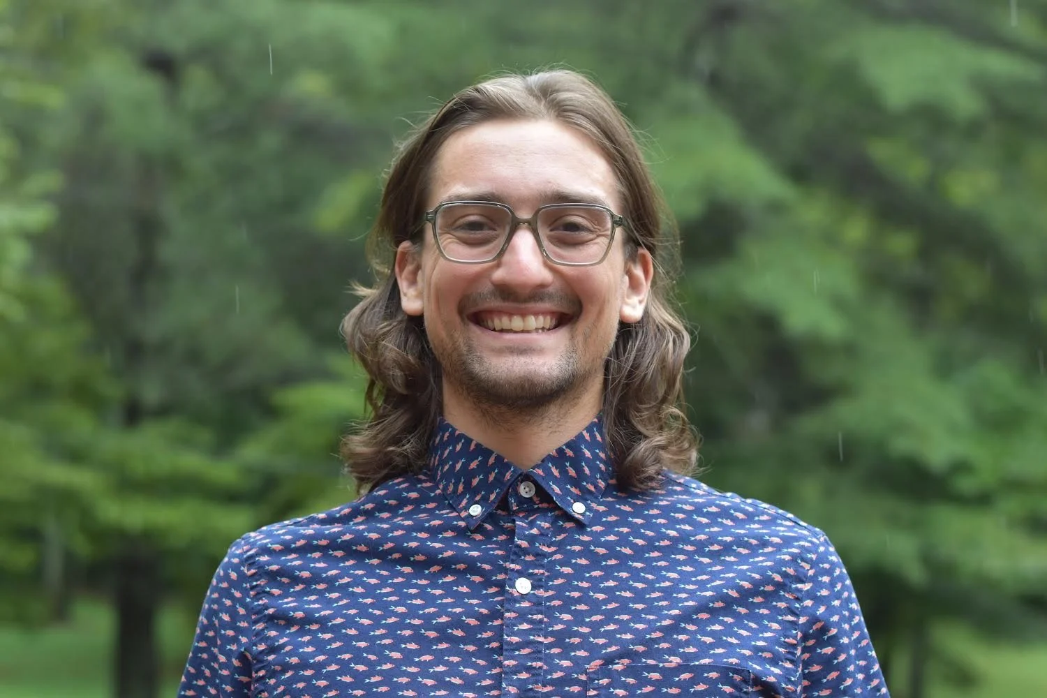 Adam stands, with dark long hair, blue shirt with gold pattern, wearing glasses
