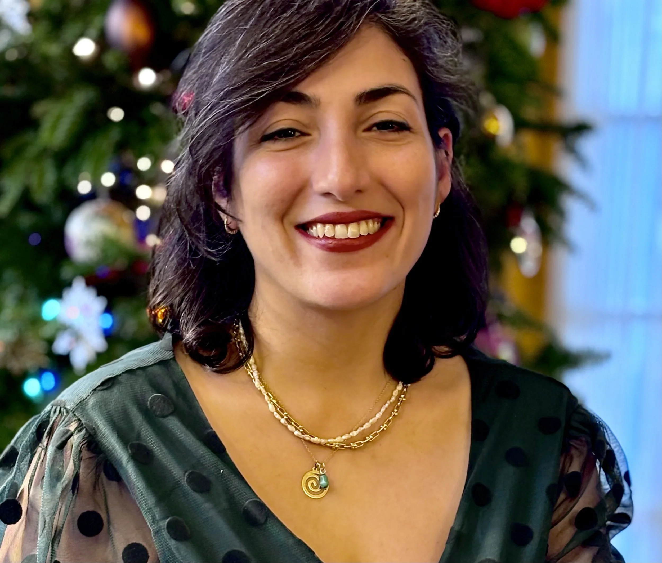 Woman smiling in front of a Christmas tree, wearing a green polka dot dress and a gold necklace.