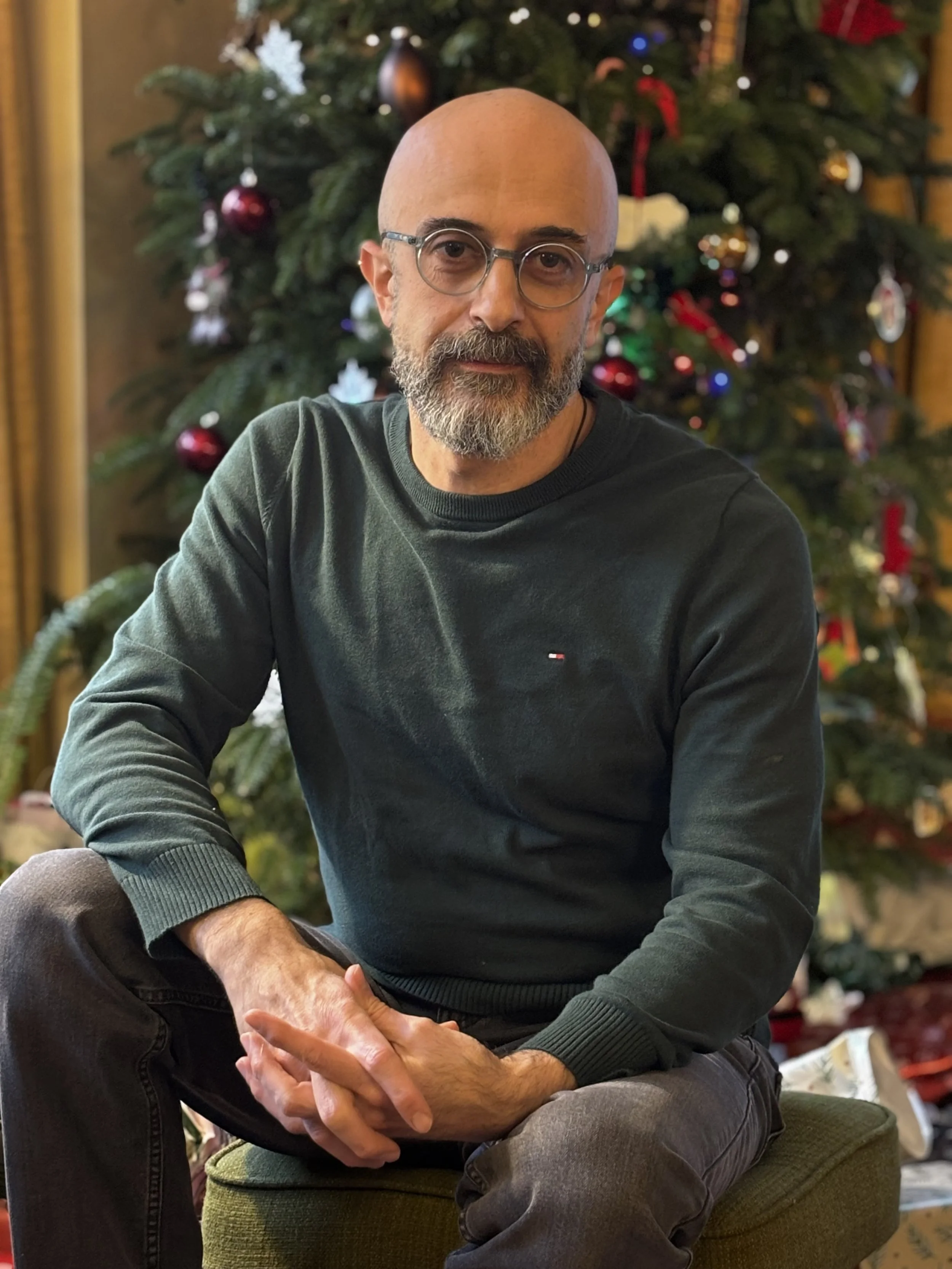 Man sitting in front of a decorated Christmas tree, wearing glasses and a dark green sweater.