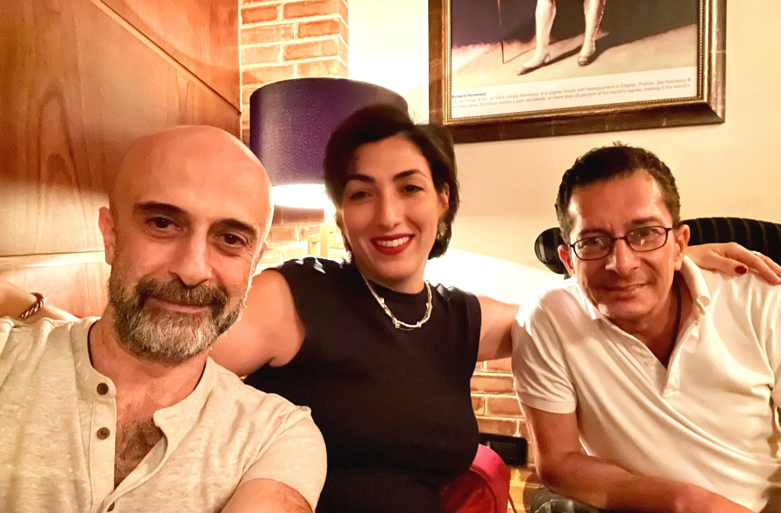 Three people sitting together indoors, smiling, with a brick wall and painting in the background.