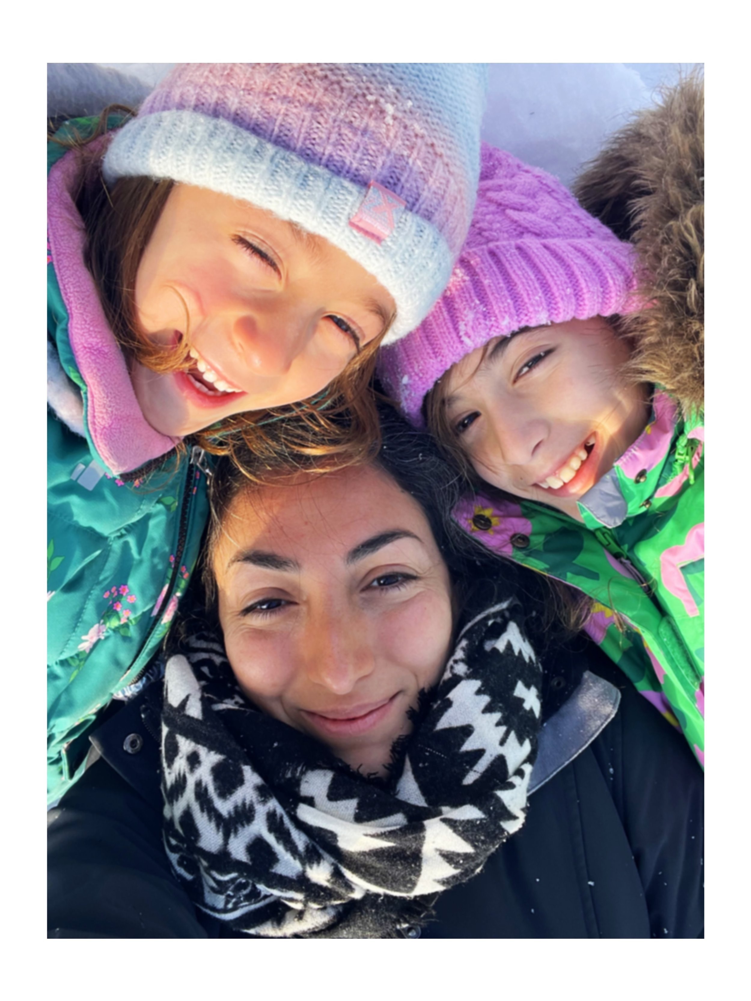 A selfie of two children in colorful winter hats and jackets, and an adult wearing a patterned scarf, all smiling and lying on the snow.