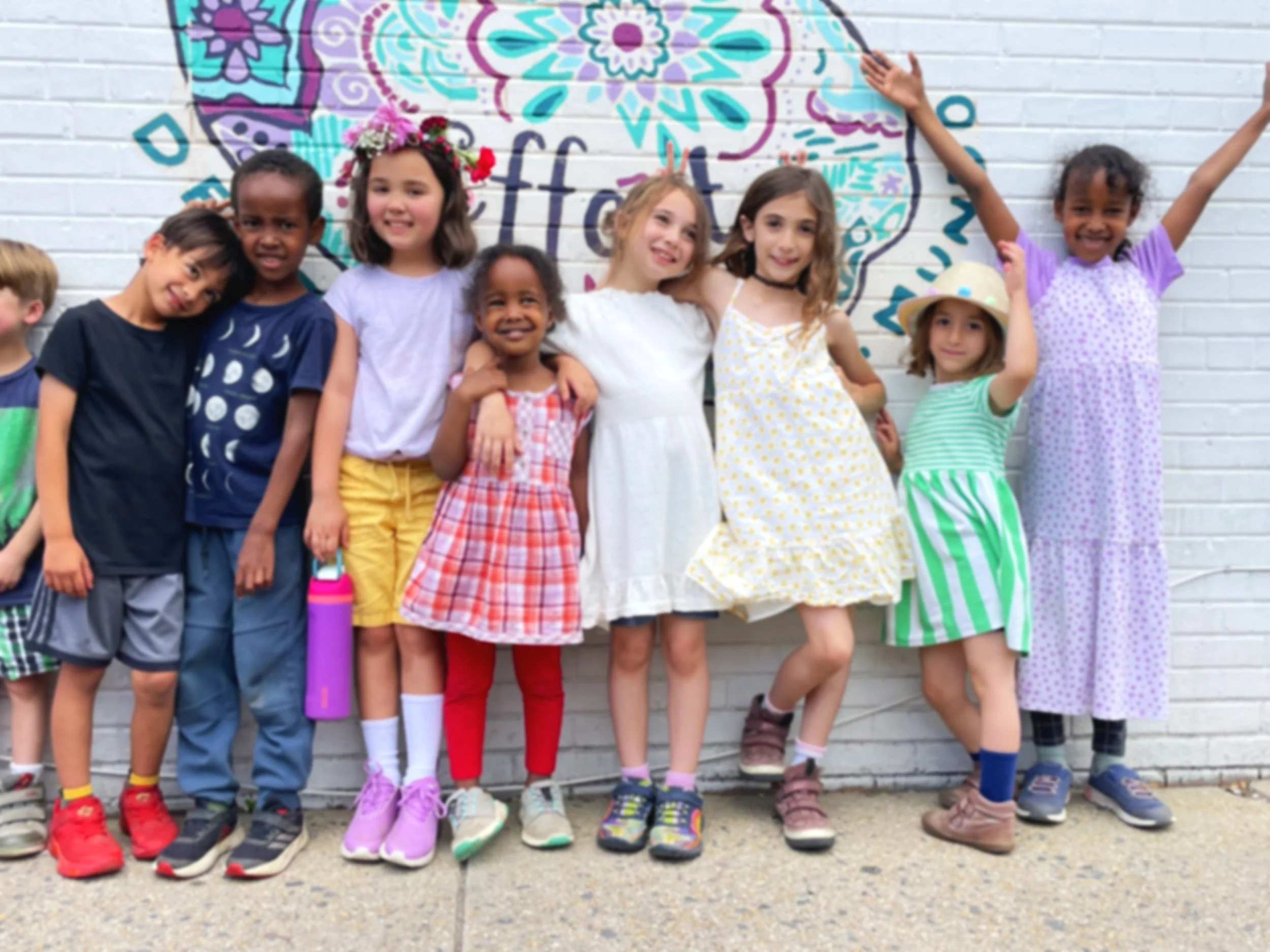 Group of children standing in front of a colorful mural painted on a white brick wall, wearing casual summer clothing and smiling.