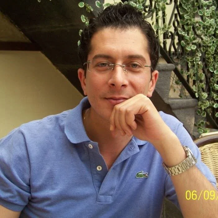 Man in blue polo shirt with glasses, sitting, hand on chin, greenery in background, date stamp on image.