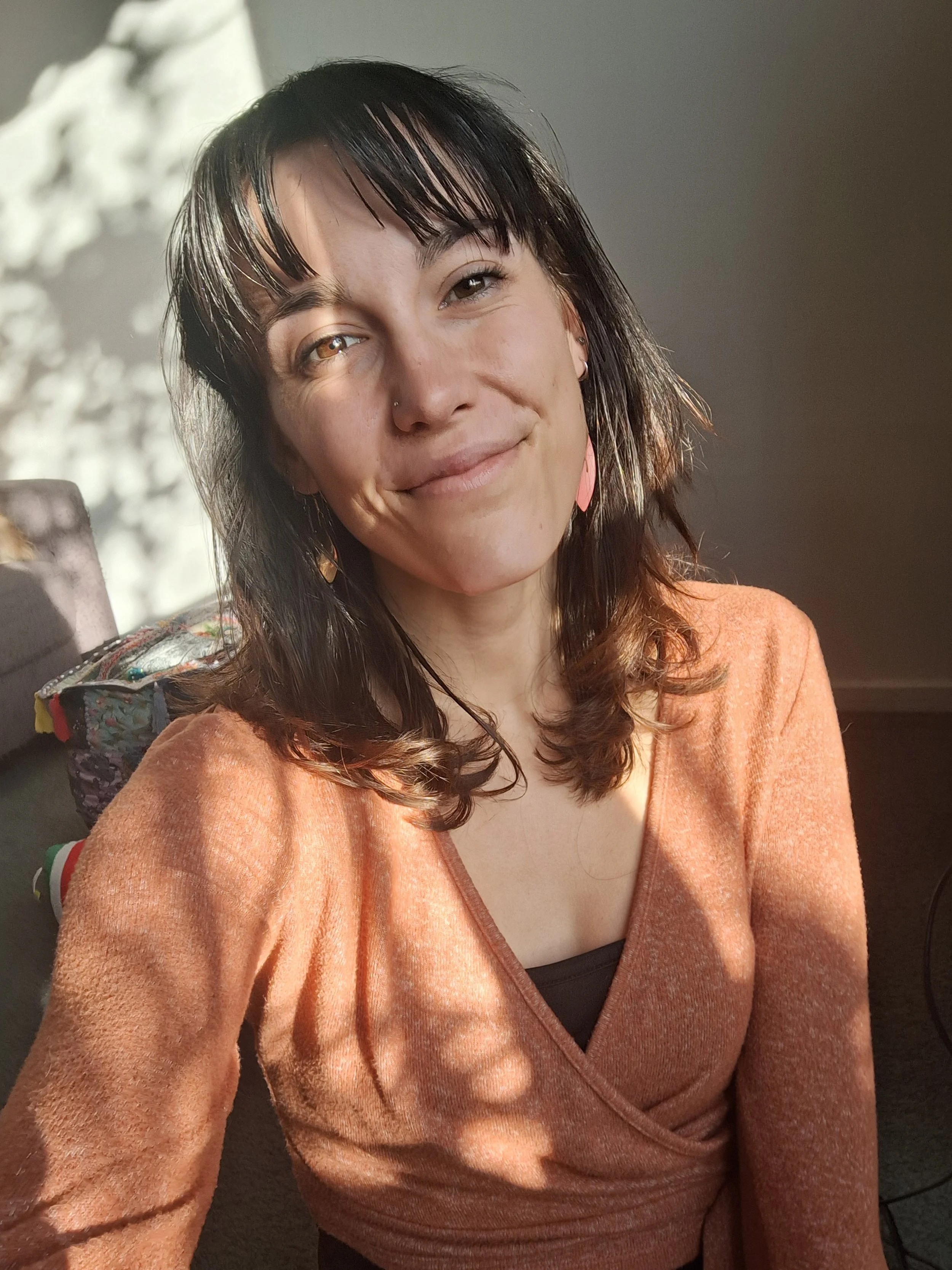 A woman with dark hair and a nose piercing smiling in natural light, wearing an orange top and pink earrings.