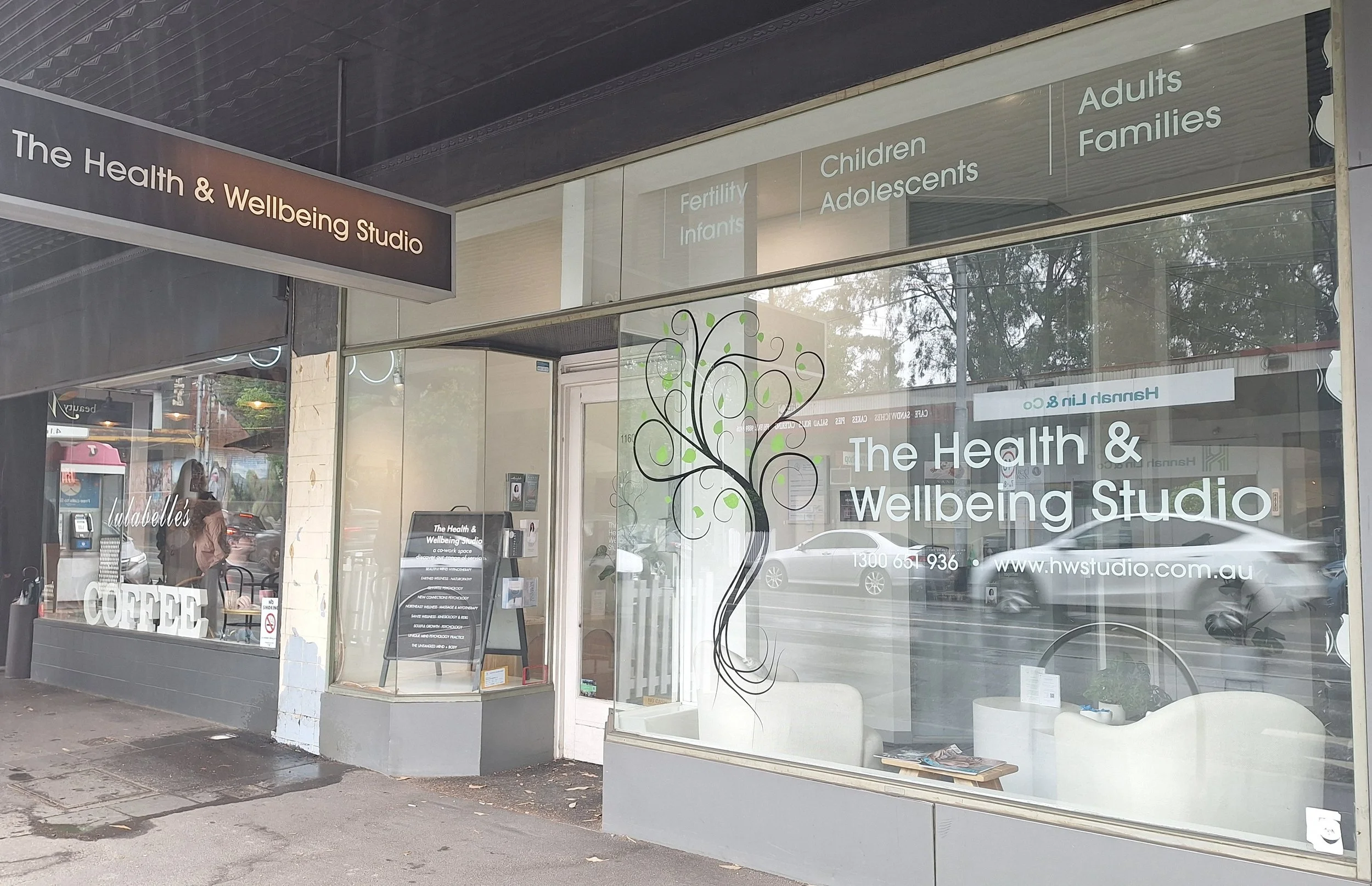 The exterior of The Health & Wellbeing Studio with large glass windows displaying the name, logo of a tree, and services for fertility, infants, children, adolescents, adults, and families. Reflection of street scene with cars and pedestrians.