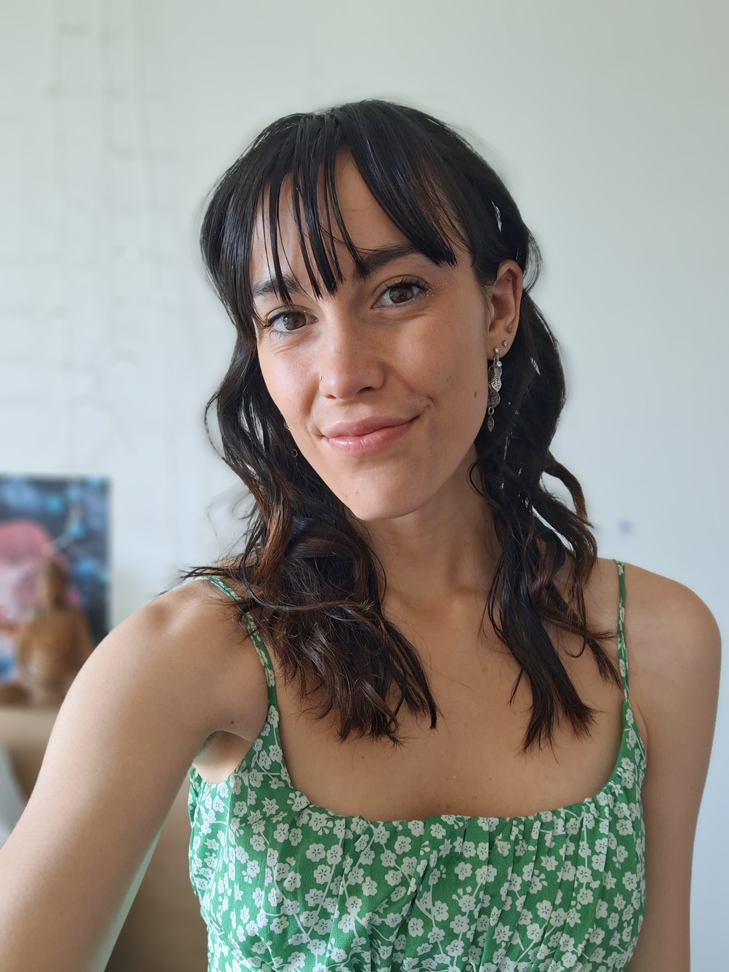 A young woman with dark hair and brown eyes smiling in a green floral dress with spaghetti straps, standing indoors with a blurred background.