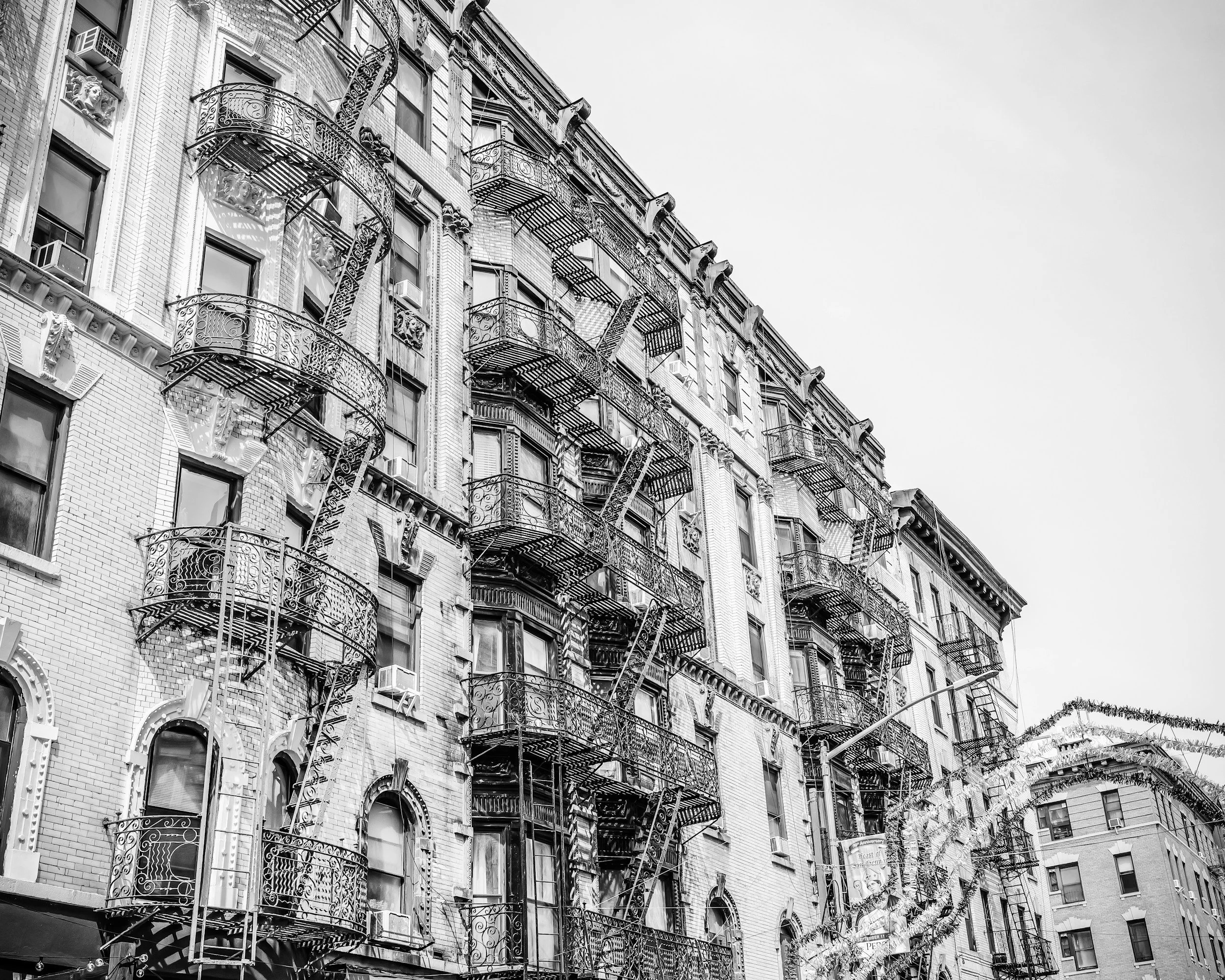 Black and white photo of a tall brick apartment building with ornate fire escapes on each floor and decorative architectural details.