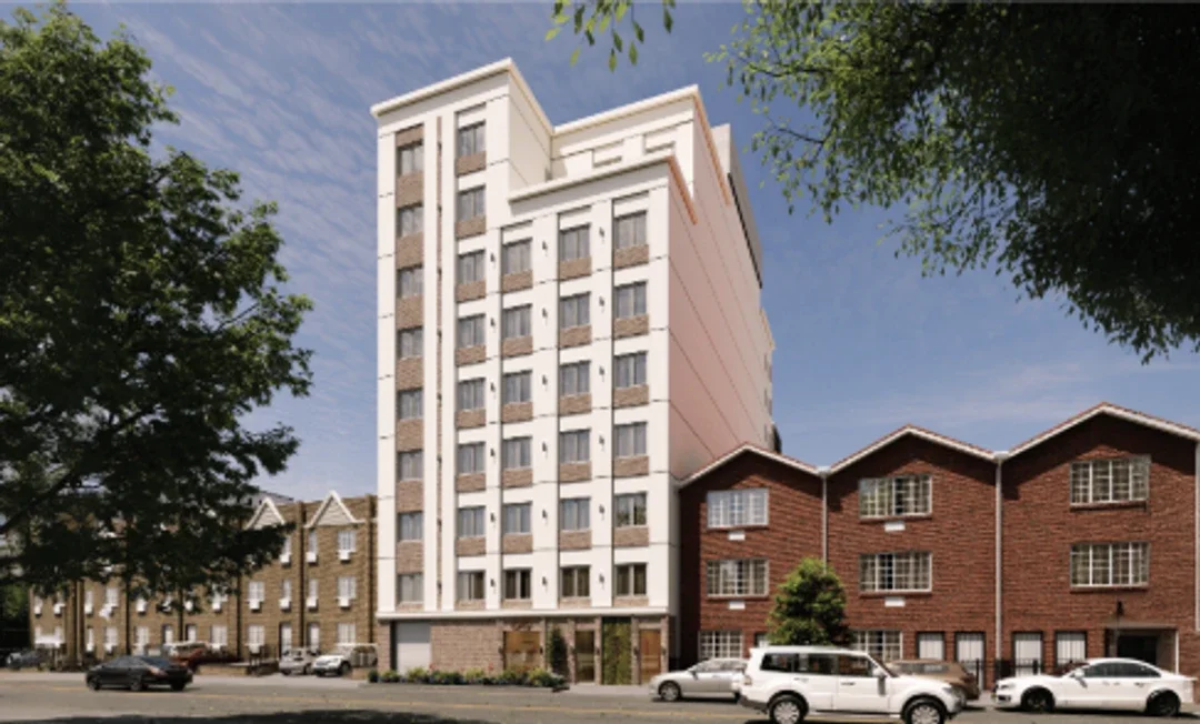 A modern multi-story residential building with a white facade and large windows, standing next to a row of red brick townhouses in an urban neighborhood, with cars parked along the street and green trees in the surroundings.