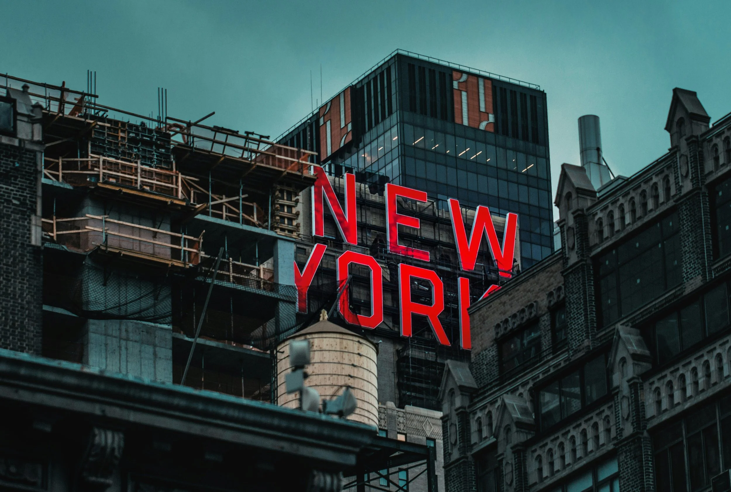 City skyscrapers under construction with a large neon sign that reads "New York" against a cloudy sky.