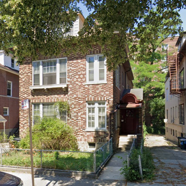 A brick multi-story residential house with a front porch, surrounded by a small yard with a fence, on a sunny day with green trees overhead.