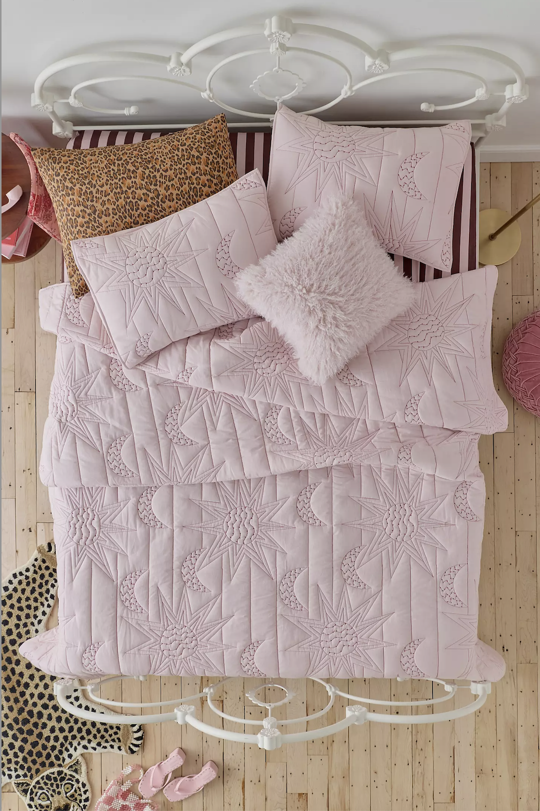 Top view of a white wrought iron bed with pink quilted bedding featuring celestial designs, three pink embroidered pillows, and a plush white pillow, in a bedroom with light wood flooring.