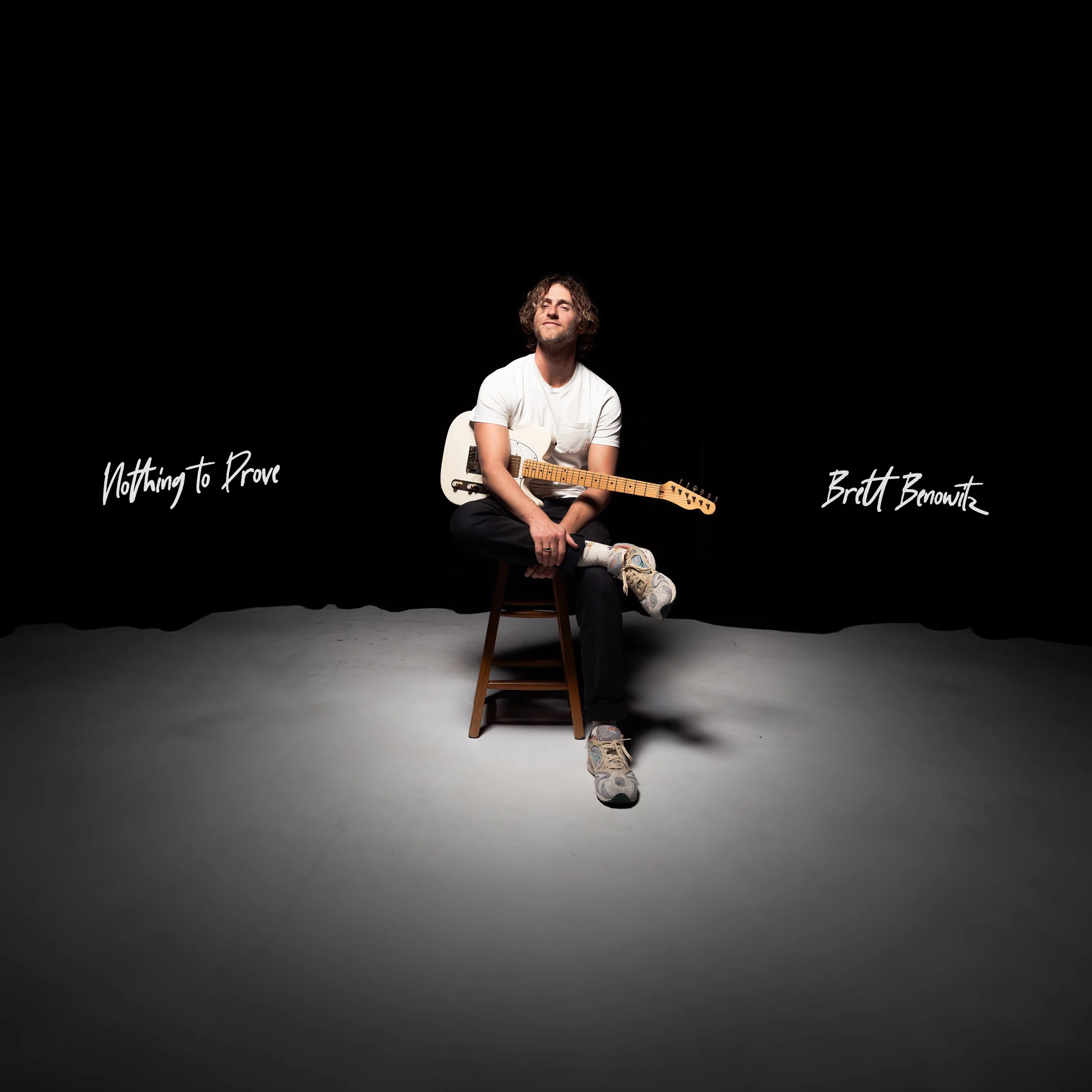 Brett Benowitz sitting on a stool under a spotlight, holding a guitar across his lap with the title ‘Nothing to Prove’ written in cursive on a dark stage backdrop.
