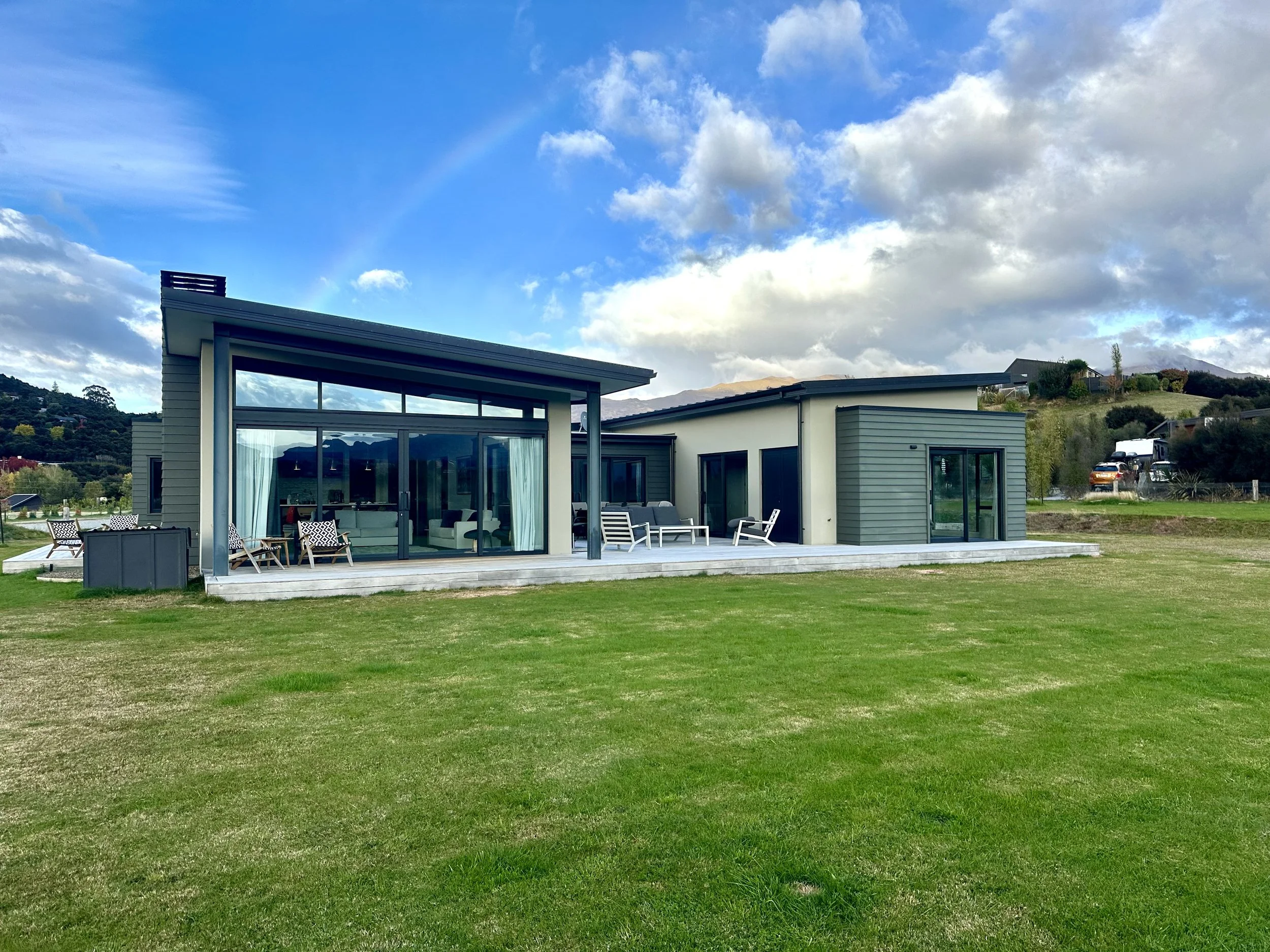 Modern house with large glass windows and a patio area, situated on a grassy lawn with hills and a partly cloudy sky in the background. - Wanaka