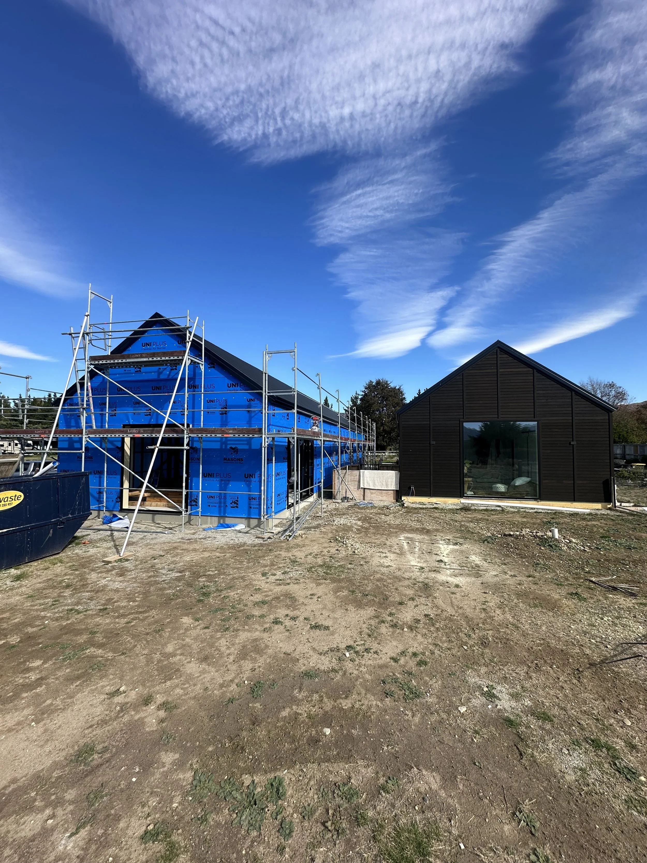Wanaka construction site with a house in progress, scaffolding and blue house wrap, next to a finished dark-colored building with a large window, on a dirt lot.
